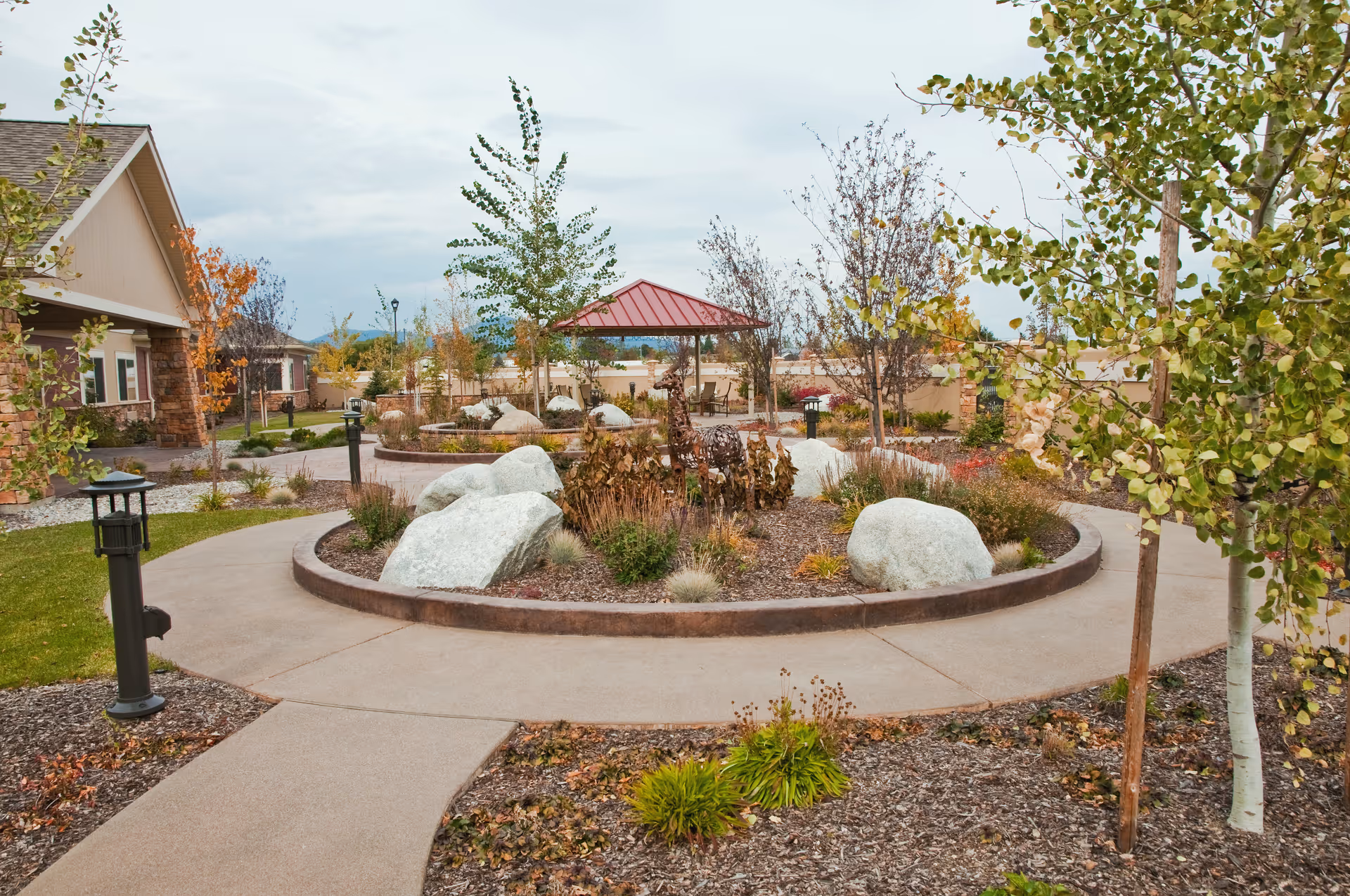 Outdoor garden area with a circular raised flower bed containing large rocks and plants, surrounded by a paved walkway. There are young trees and shrubs around the garden, a small gazebo with a red roof in the background, and a building with stone and beige siding on the left side.