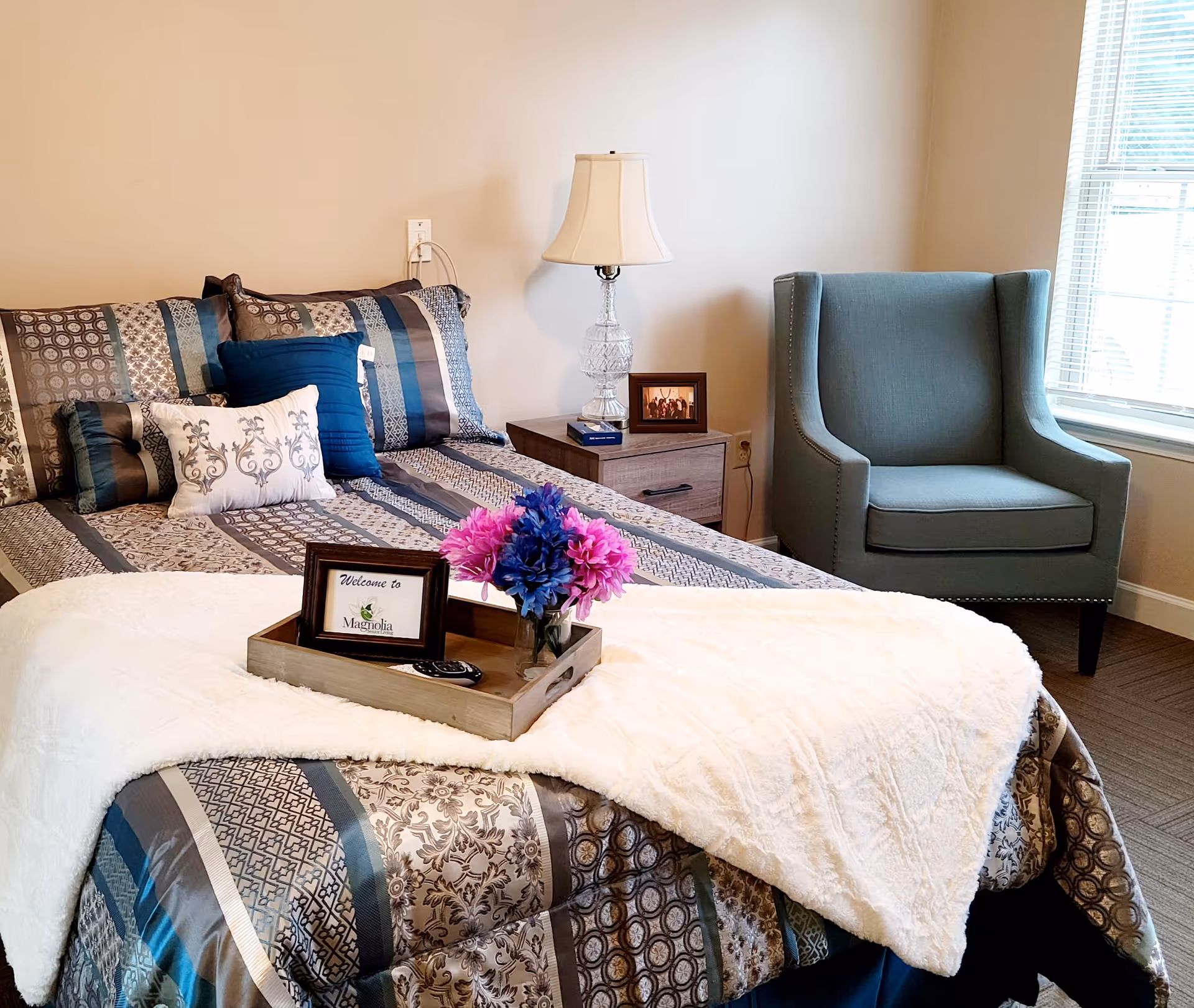 A neatly made bed with patterned bedding and multiple pillows in a senior living facility bedroom. A wooden tray on the bed holds a small framed sign that says 'Welcome to Magnolia' and a vase with purple and pink flowers. Next to the bed is a nightstand with a clear glass lamp and a small framed photo. A blue upholstered armchair is positioned near a window with blinds.