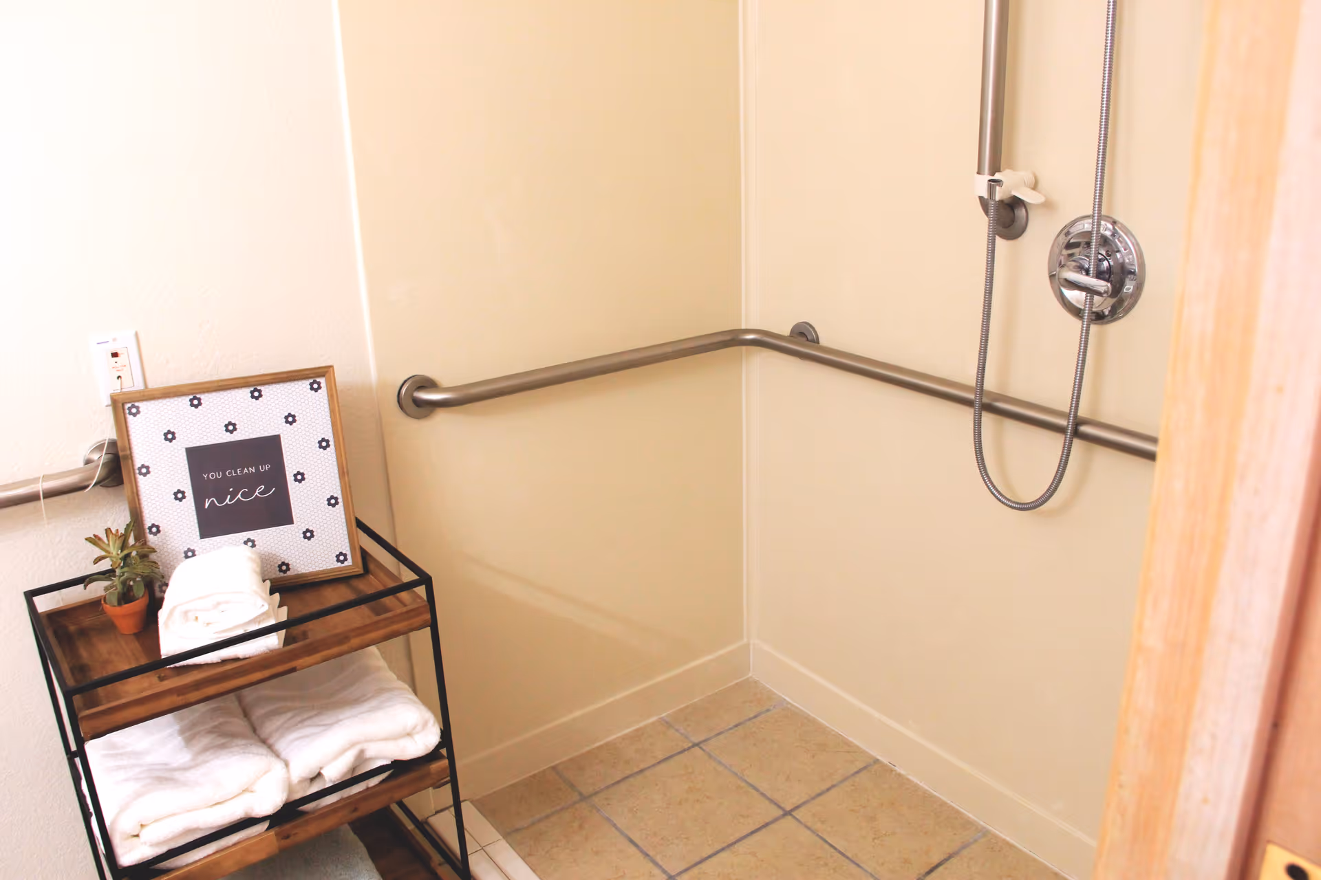 Accessible shower area with beige walls and tiled floor, equipped with a handheld showerhead and horizontal grab bars. Next to the shower is a wooden shelf holding folded white towels, a small potted plant, and a framed sign that reads 'You clean up nice.'
