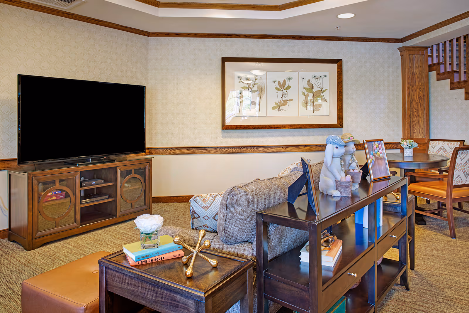 A cozy living room area with a large flat-screen TV on a wooden cabinet, a gray upholstered sofa, a wooden side table with books and decorative items, and a wooden console table with framed photos and bunny figurines. In the background, there is a round table with chairs and a staircase with wooden railings. The walls have patterned wallpaper and a framed botanical artwork.