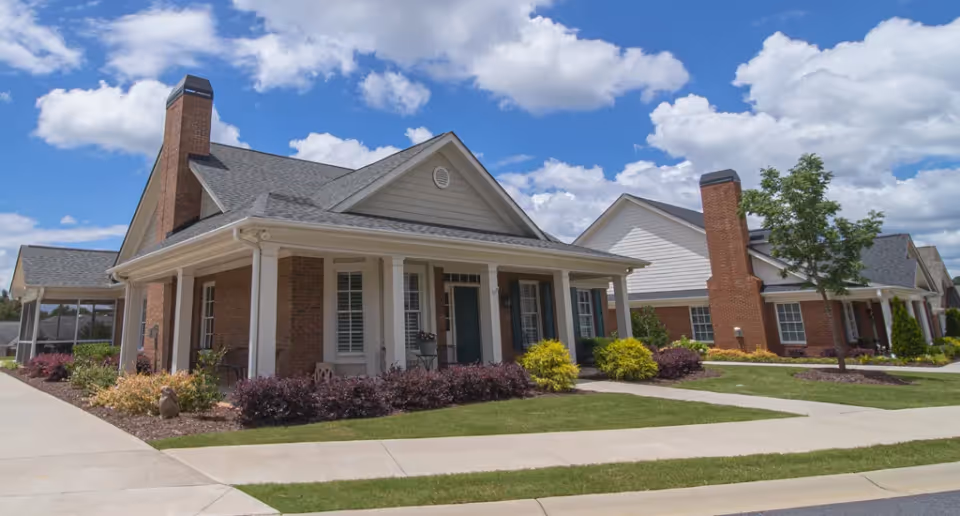 Exterior view of a residential-style building with brick walls, white columns, and a covered porch under a partly cloudy blue sky. The building is surrounded by landscaped bushes, a tree, and a well-maintained lawn with concrete walkways.