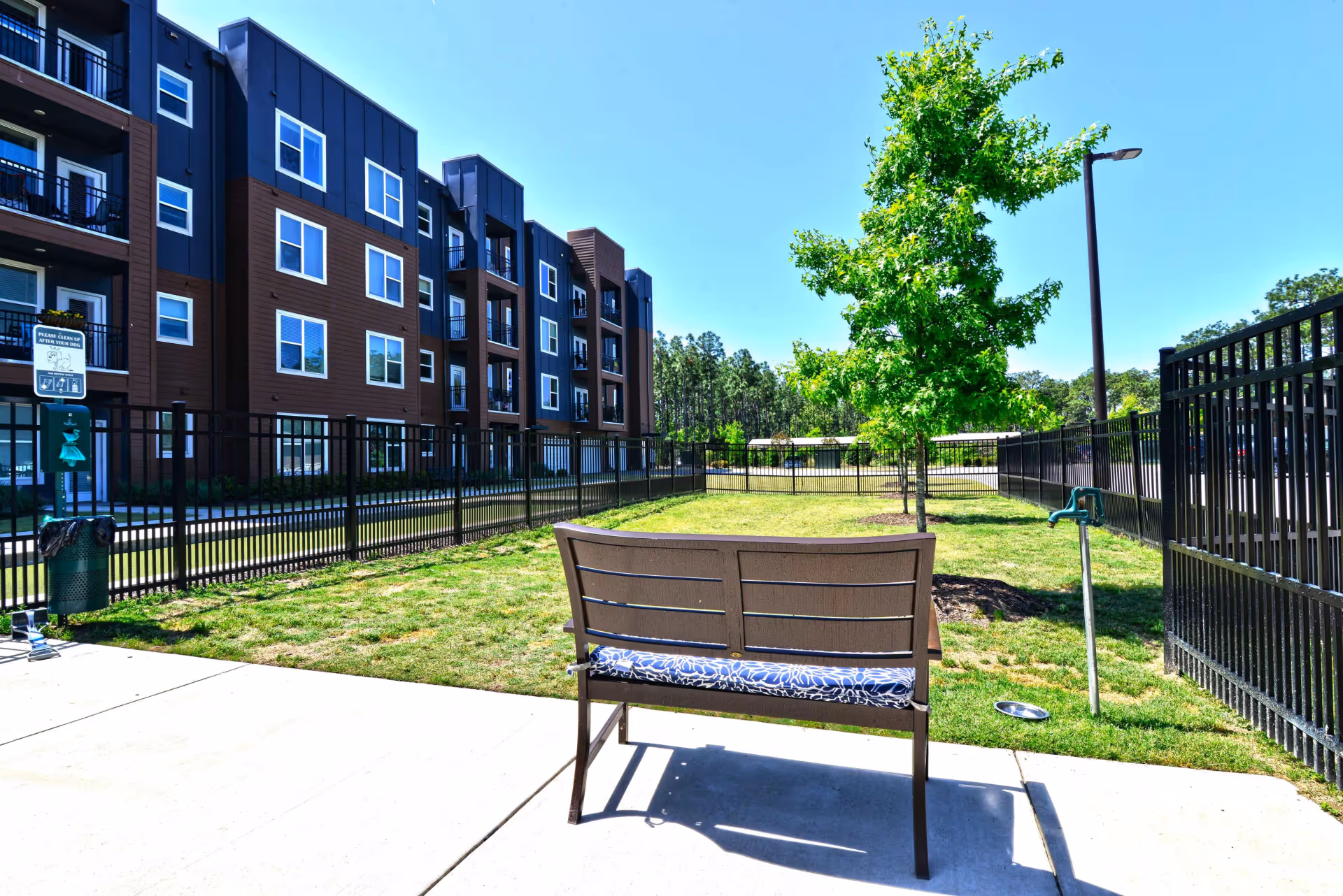 Outdoor area of a senior living facility with a bench facing a grassy fenced-in dog park, a tree, a water faucet, and a multi-story building in the background under a clear blue sky.