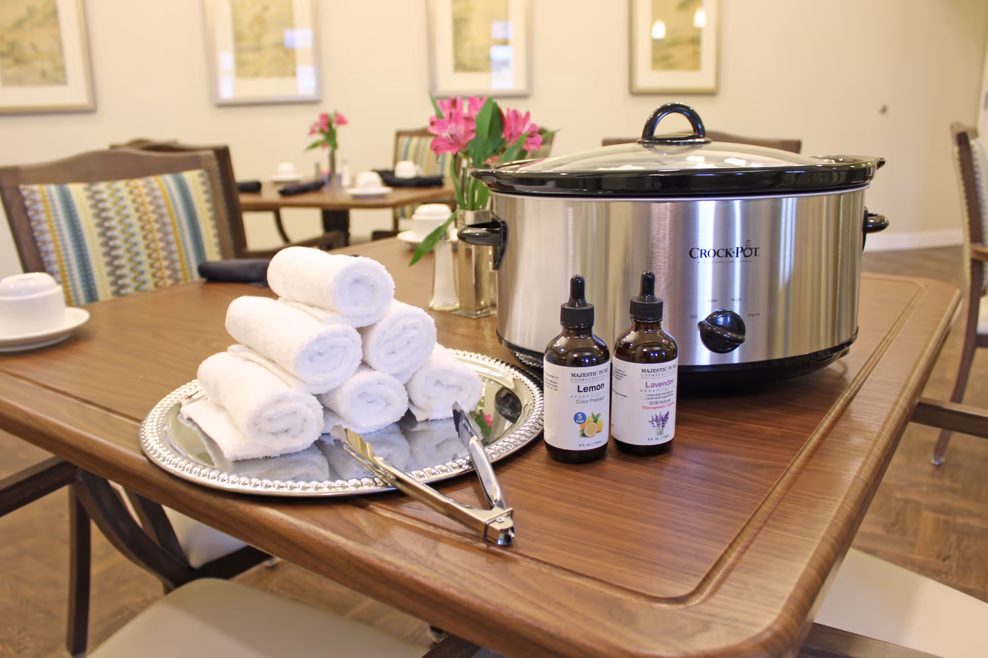 A wooden table with a silver tray holding six rolled white towels and a pair of tongs, next to a large stainless steel Crock-Pot slow cooker and two small bottles of essential oils labeled Lemon and Lavender. In the background, there are chairs with patterned cushions and framed artwork on the wall.