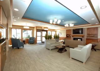 A spacious and well-lit living room area in an assisted living facility featuring light-colored carpet, two white sofas, a coffee table with a flower arrangement, a fireplace with a mounted TV above it, wooden cabinetry, and several armchairs near large windows allowing natural light to enter.