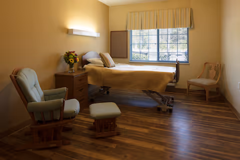 A simple, well-lit bedroom in a senior living facility featuring a hospital-style adjustable bed with striped bedding, a wooden nightstand with a vase of flowers, a cushioned rocking chair with a matching footrest, and a wooden chair near a large window with blinds and a valance.