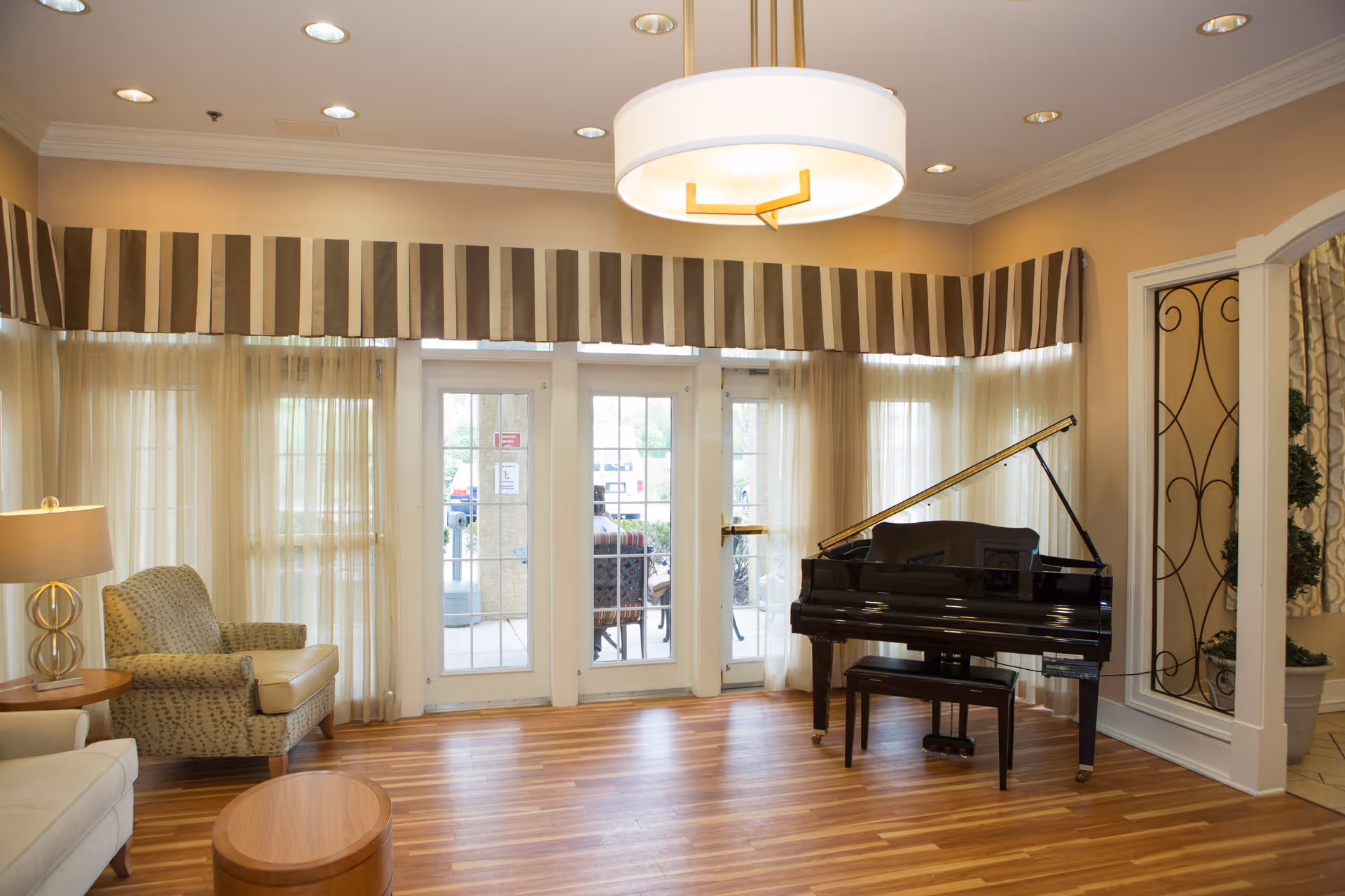 A bright and inviting living room area with wooden flooring, a black grand piano with a matching bench, a beige armchair, a small round wooden table, and a lamp with a circular base. Large windows and glass doors with sheer curtains and striped valances allow natural light to fill the room.