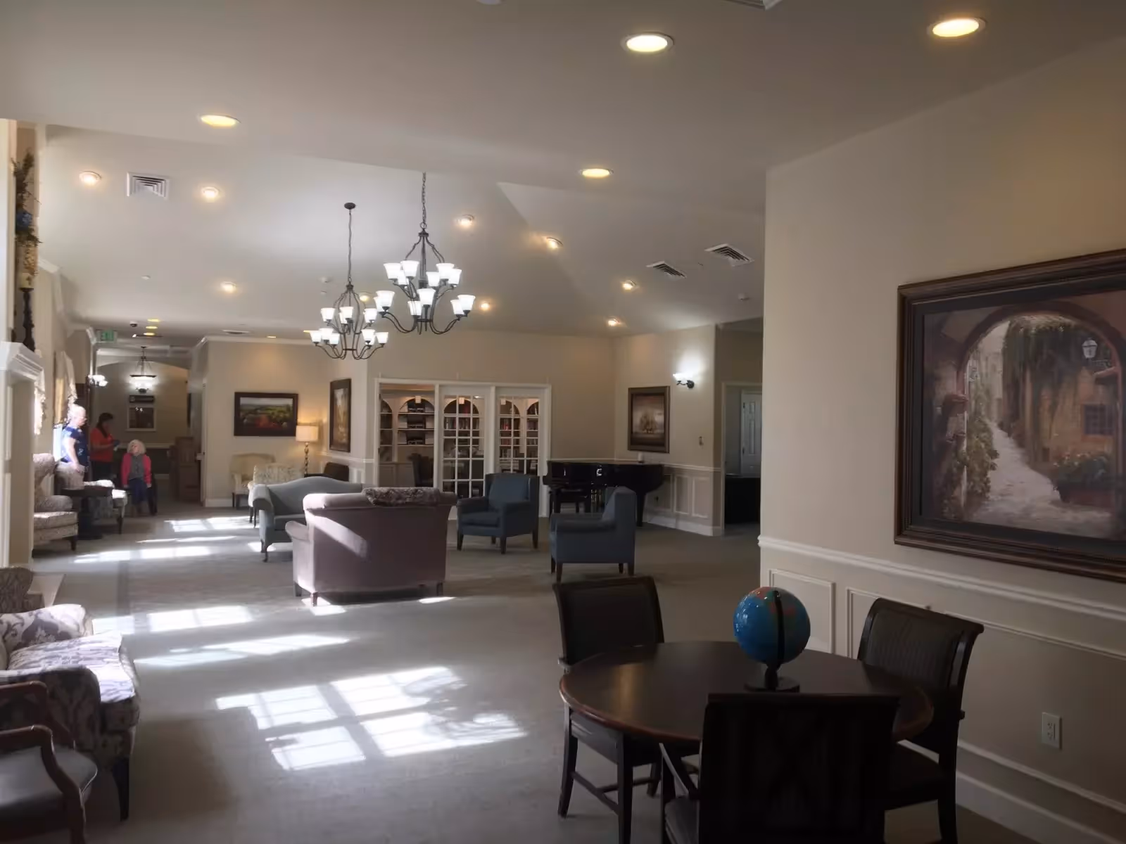 A spacious and well-lit common area in an assisted living facility featuring multiple seating arrangements including armchairs and sofas. There are chandeliers hanging from the ceiling, framed artwork on the walls, and a round table with chairs and a globe on it in the foreground. Several people are visible in the background near the windows.