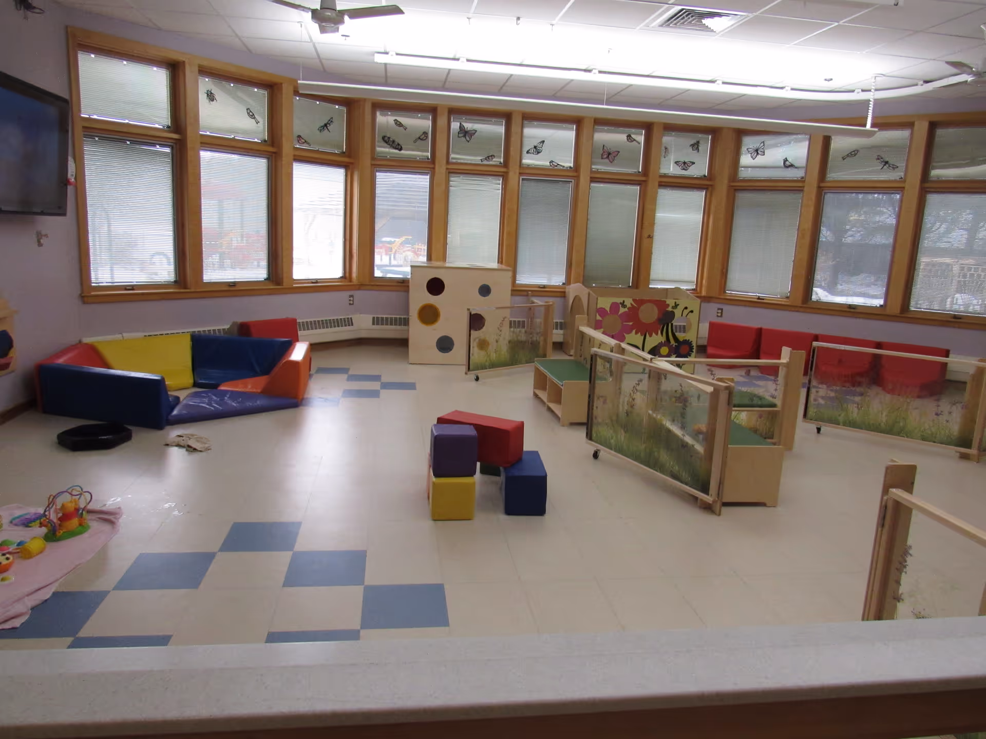 A brightly lit indoor playroom with large windows featuring butterfly decals. The room contains colorful padded seating, small soft blocks, a play mat with toys, and wooden play structures with nature-themed panels. The floor has a checkered pattern with blue and white tiles.