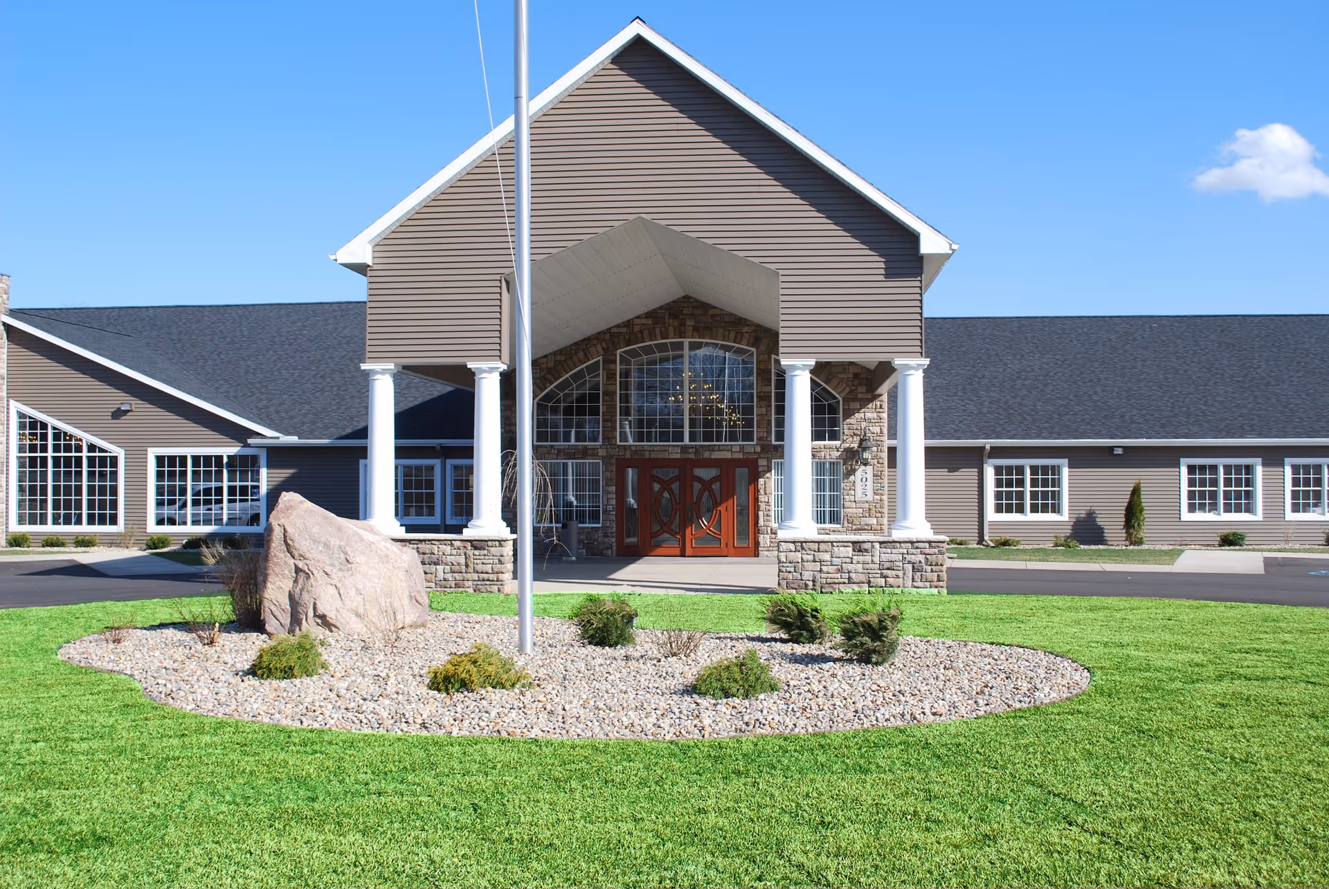 Front exterior view of a single-story assisted living facility building with a peaked roof, large windows, and a covered entrance supported by white columns. There is a landscaped area with rocks, shrubs, and a large rock in front of the building, along with a flagpole. The sky is clear and blue.