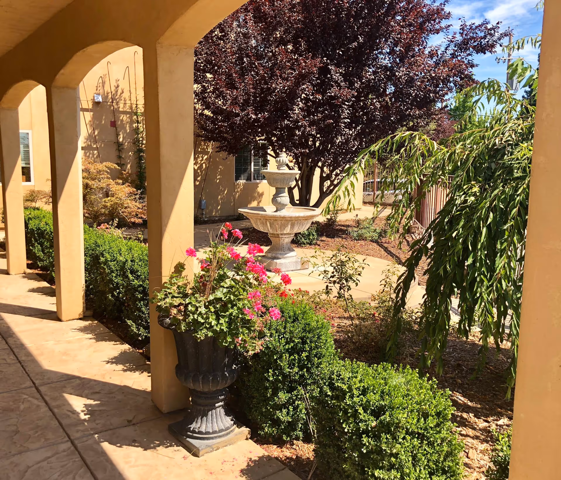 Sunlit courtyard with arched columns, a planter of pink flowers, trimmed hedges and a central stone fountain.