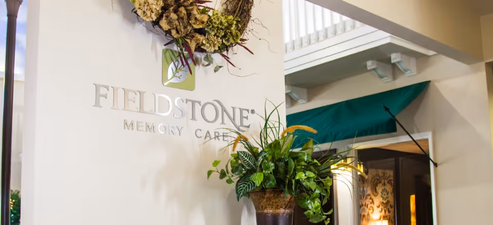 Interior view of a memory care facility with a white wall displaying the Fieldstone Memory Care logo, a decorative wreath above the logo, and a large potted plant in front of the wall. In the background, there is a green awning and part of a room with patterned wallpaper and warm lighting.