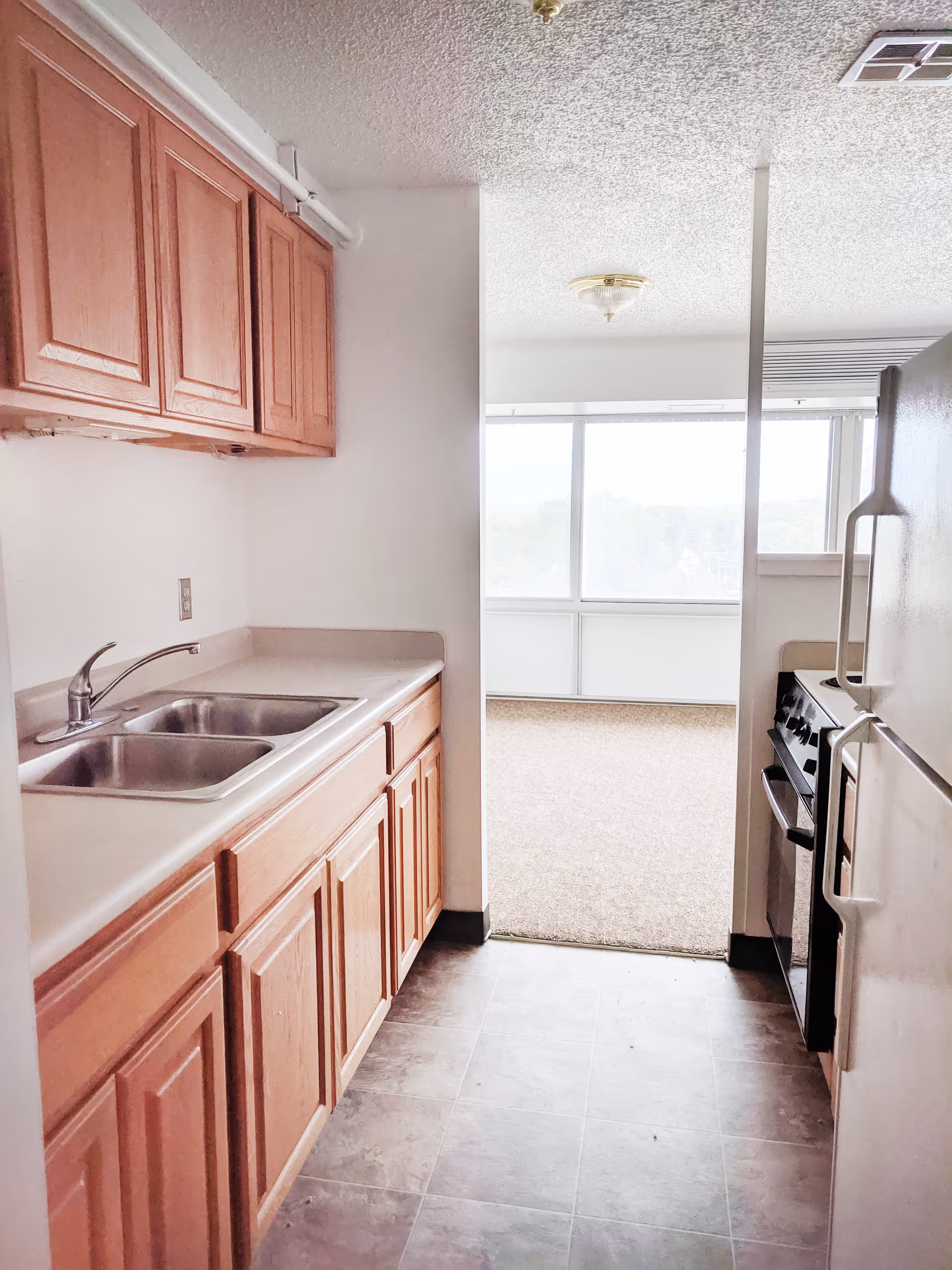Small galley kitchen with wooden cabinets, a double sink, stove and refrigerator opening into a bright windowed living area.
