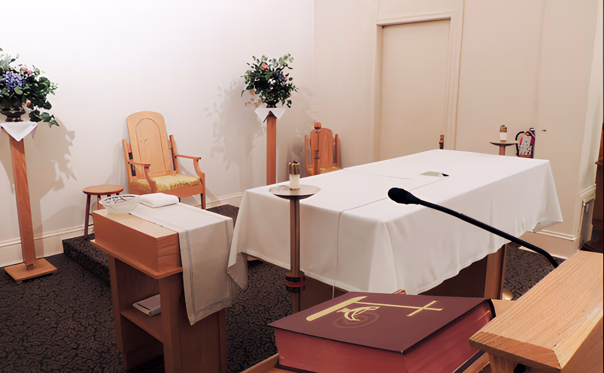 Small chapel interior with a white-covered altar, lectern with a microphone and Bible, wooden chairs and floral arrangements.
