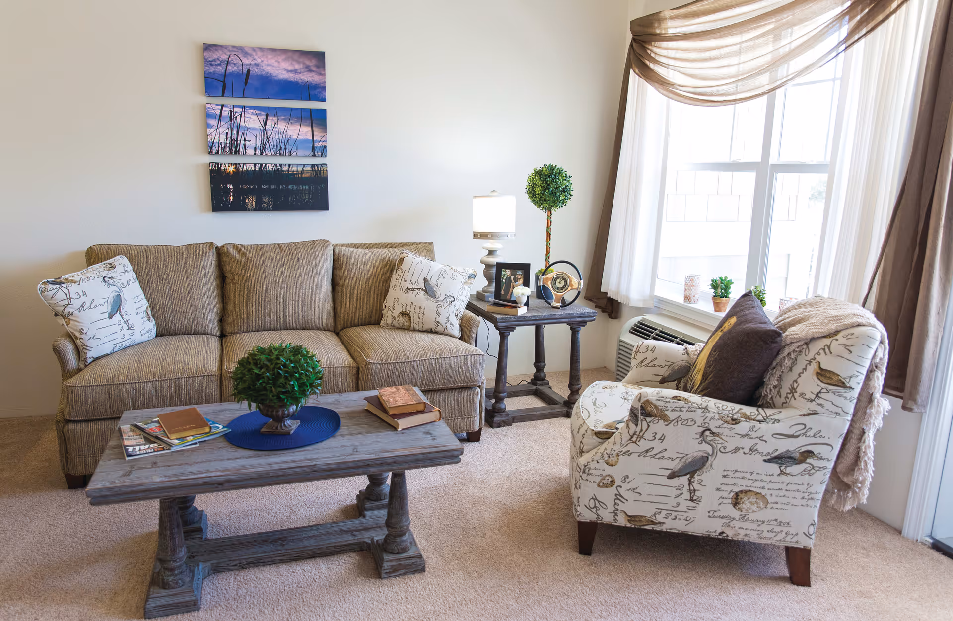 A cozy living room with a beige sofa adorned with patterned pillows, a wooden coffee table with books and a small plant, a side table with a lamp and decorative items, and a patterned armchair near a large window with sheer and brown curtains.