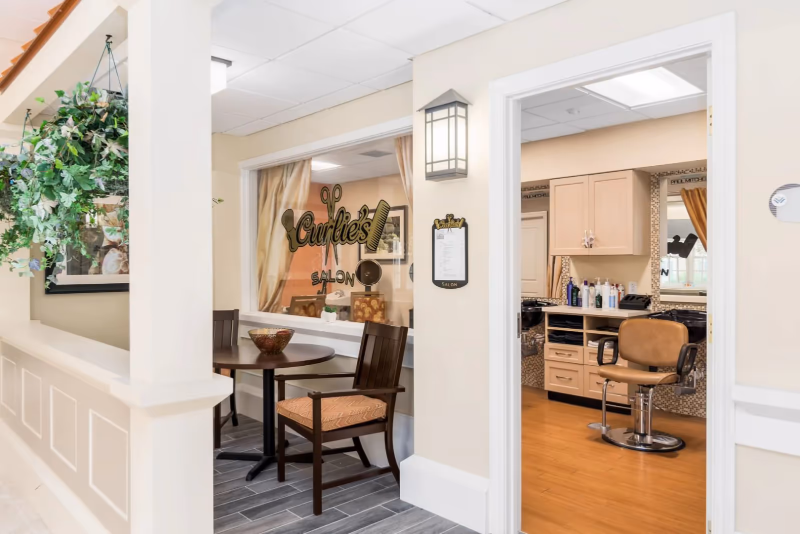 Interior of a salon area showing a small table and chairs by a window with 'Curlies Salon' lettering and an open doorway to a salon chair and cabinetry.