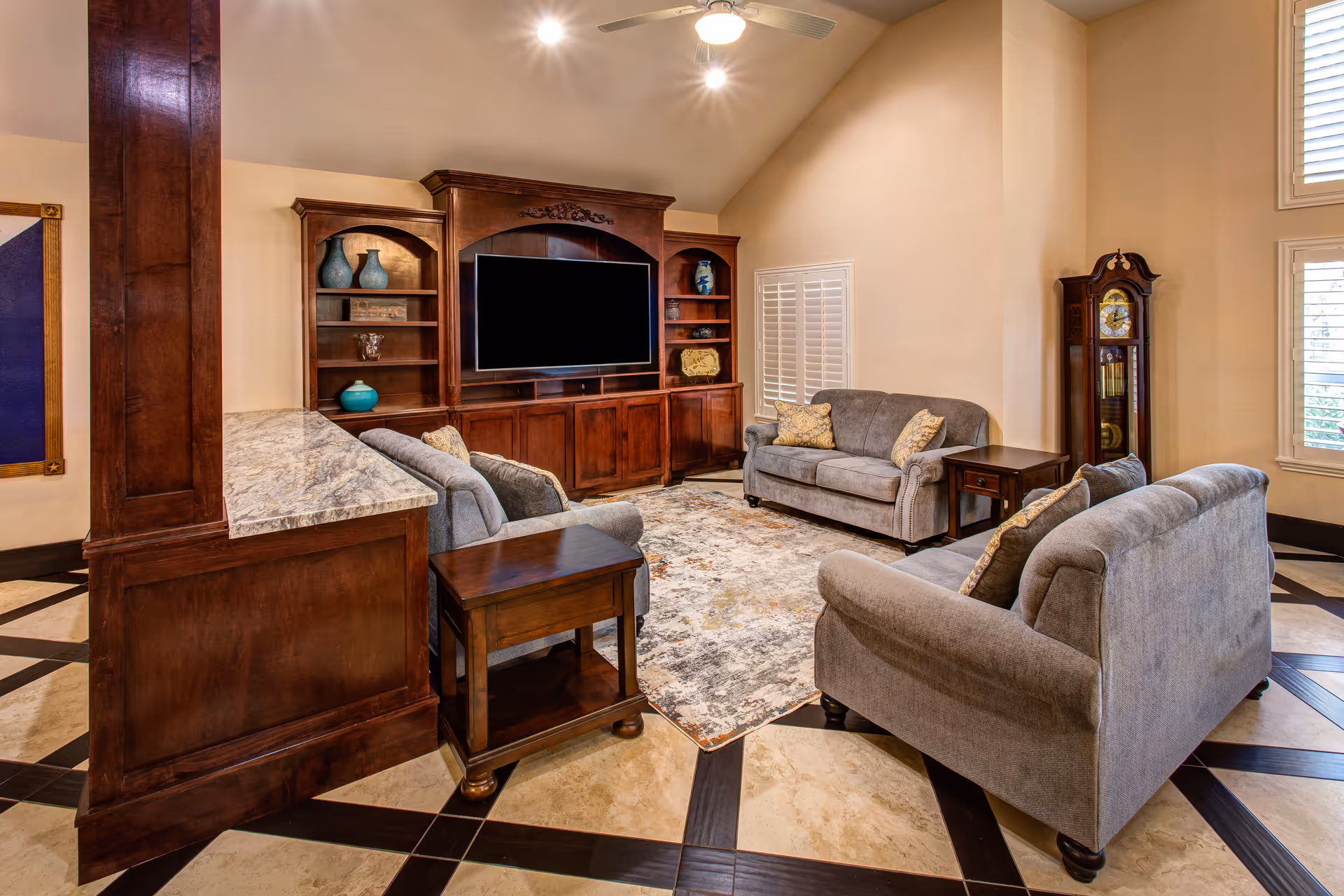 A cozy living room with three gray upholstered sofas arranged around a patterned area rug. There is a large wooden entertainment center with a flat-screen TV and decorative vases. The room features a tall ceiling with recessed lighting and a ceiling fan. A grandfather clock stands near the window with white plantation shutters.