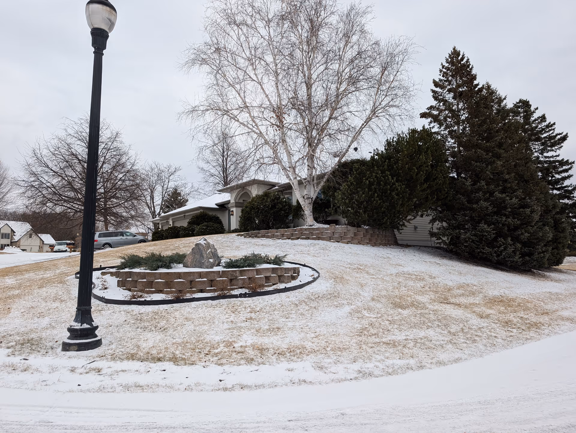 Front exterior of a single-story home with a snowy lawn, trees, a stone planter and a lamppost.
