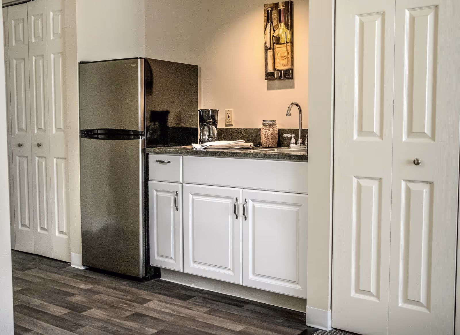 A small kitchen area with a stainless steel refrigerator, white cabinets with silver handles, a countertop with a coffee maker, a jar filled with corks, and a sink with a faucet. There is a painting of wine bottles on the wall above the countertop. The floor has a wood-like finish, and there are white paneled closet doors on either side of the kitchen area.