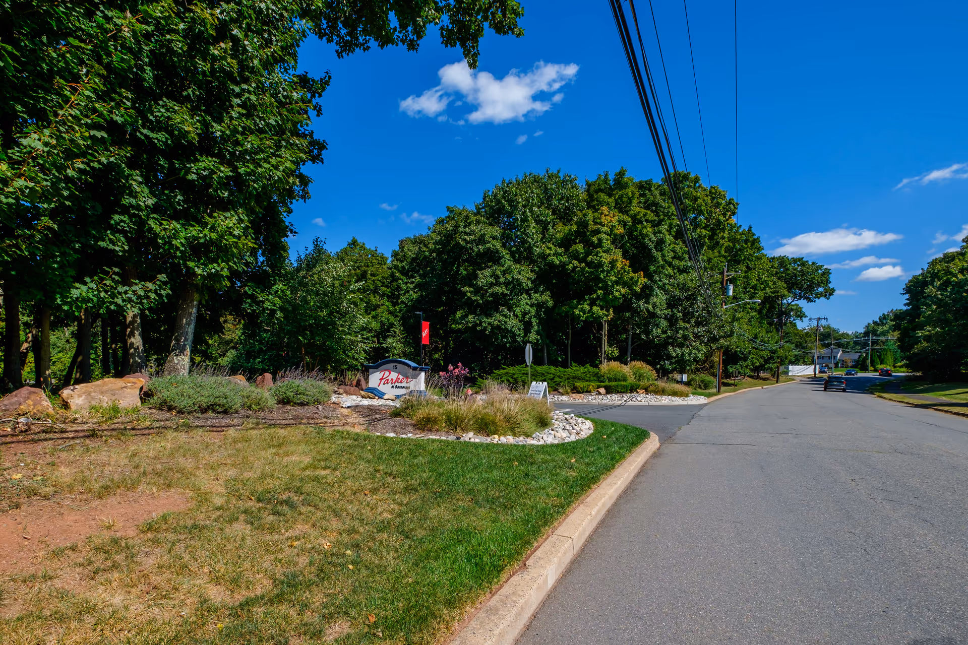 A clear sunny day view of a road lined with trees and greenery on the left side. There is a landscaped area with bushes, rocks, and a sign that reads 'Parker At Somerset'. Power lines run along the right side of the road, and a few cars are visible in the distance.