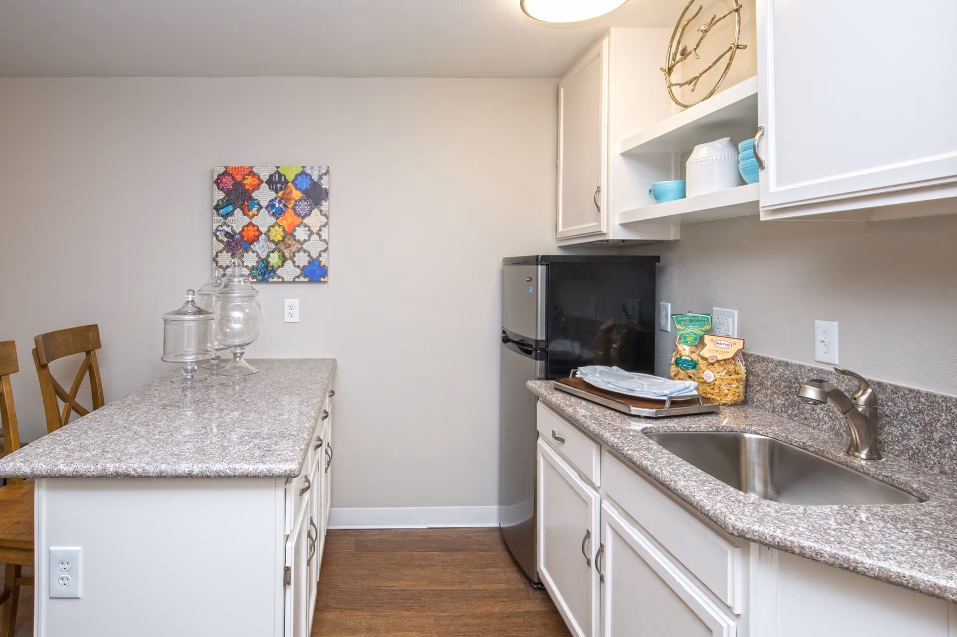 A small kitchen area with white cabinets, a granite countertop, a stainless steel sink, and a mini refrigerator. There are two wooden chairs at a granite countertop island, decorative jars on the island, and a colorful patterned wall art hanging on the wall.