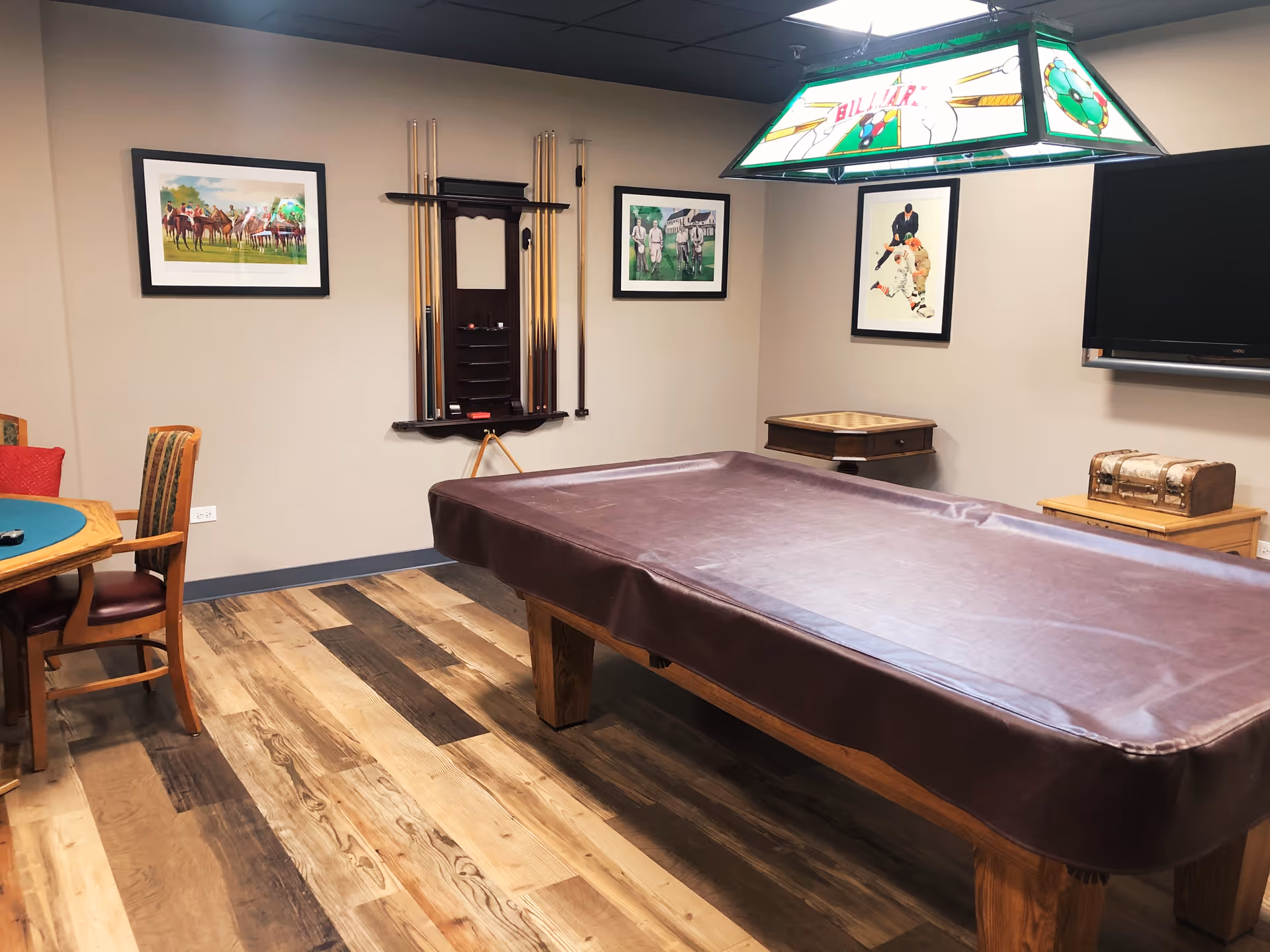 A recreational room with a covered pool table in the center, a poker table with chairs on the left, a wall-mounted rack holding pool cues, framed artwork on the walls, a small table with a chessboard, a treasure chest decoration, and a flat-screen TV mounted on the wall.