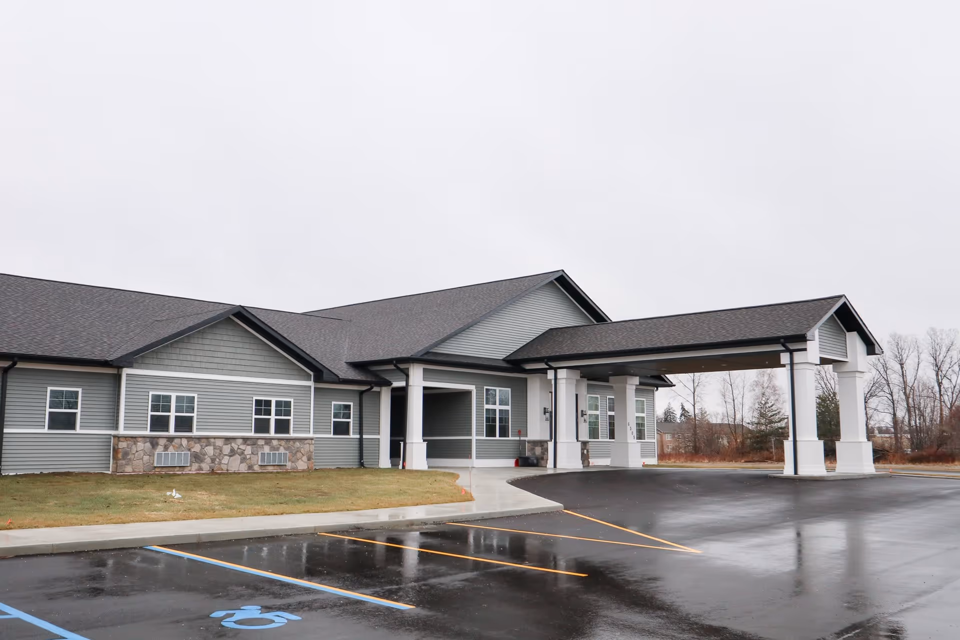 Exterior view of a single-story building with gray siding and stone accents, featuring a covered entrance supported by white columns. The parking lot in front has marked spaces including a handicapped parking spot. The sky is overcast and the surrounding area has some trees and grass.