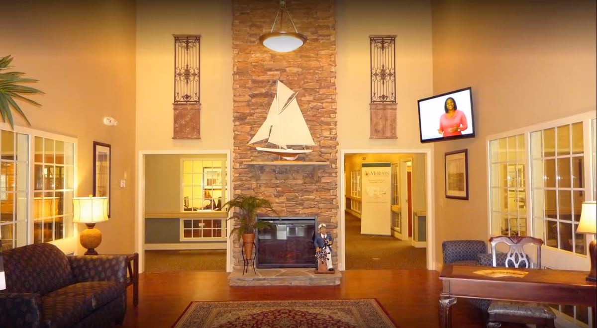 Senior living common room with a stone fireplace topped by a decorative sailboat, seating areas, lamps and a wall-mounted TV.