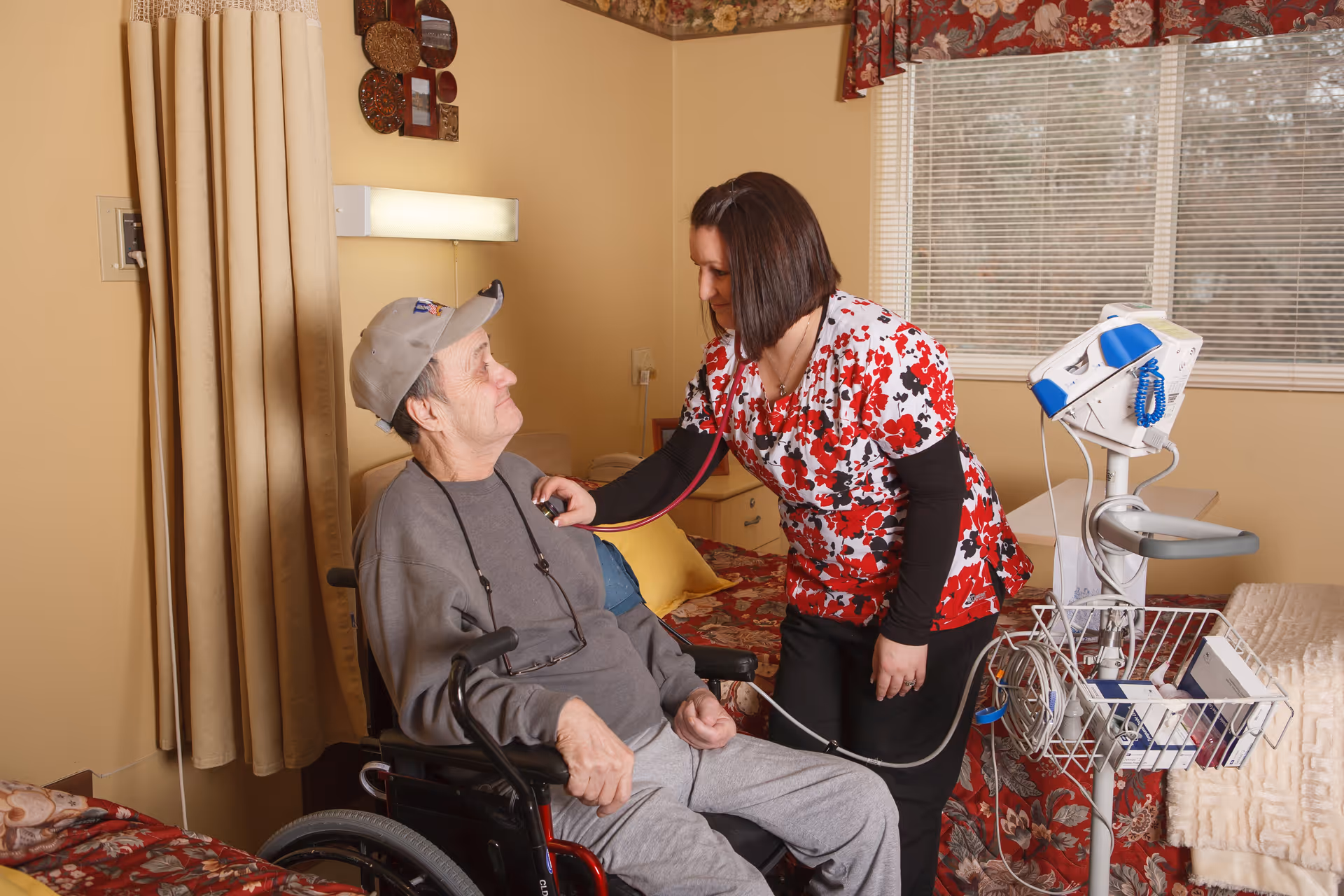 A healthcare worker in a floral scrub top is using a stethoscope to check the heartbeat of an elderly man sitting in a wheelchair inside a room with beige walls, a bed with floral bedding, and a medical device on a stand nearby.
