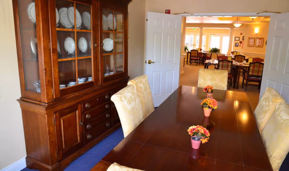 Dining room with a wooden china cabinet, a long polished table with small flower centerpieces, and upholstered chairs opening into a larger dining area.