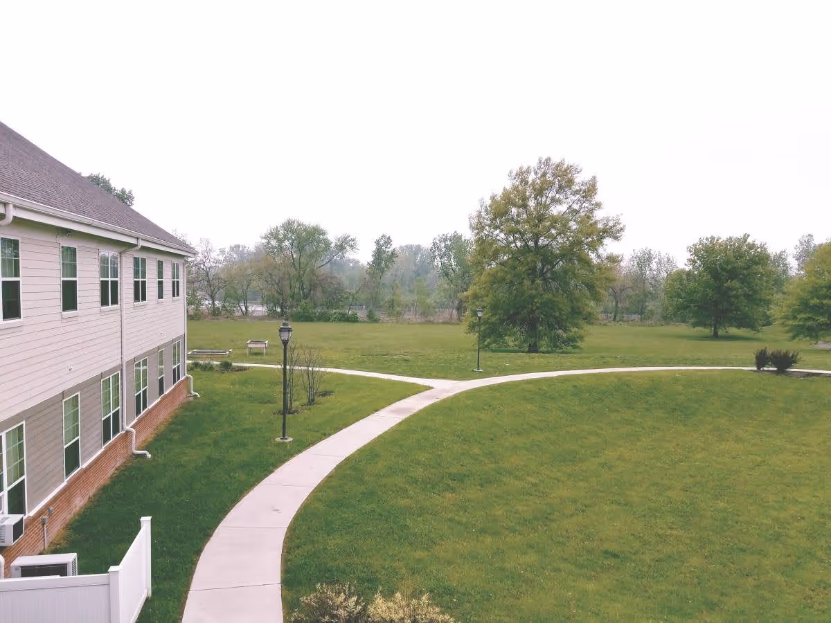 Outdoor view of a senior living facility named Cranes Landing showing a curved concrete pathway, green grass, several trees, and part of a two-story building with multiple windows.