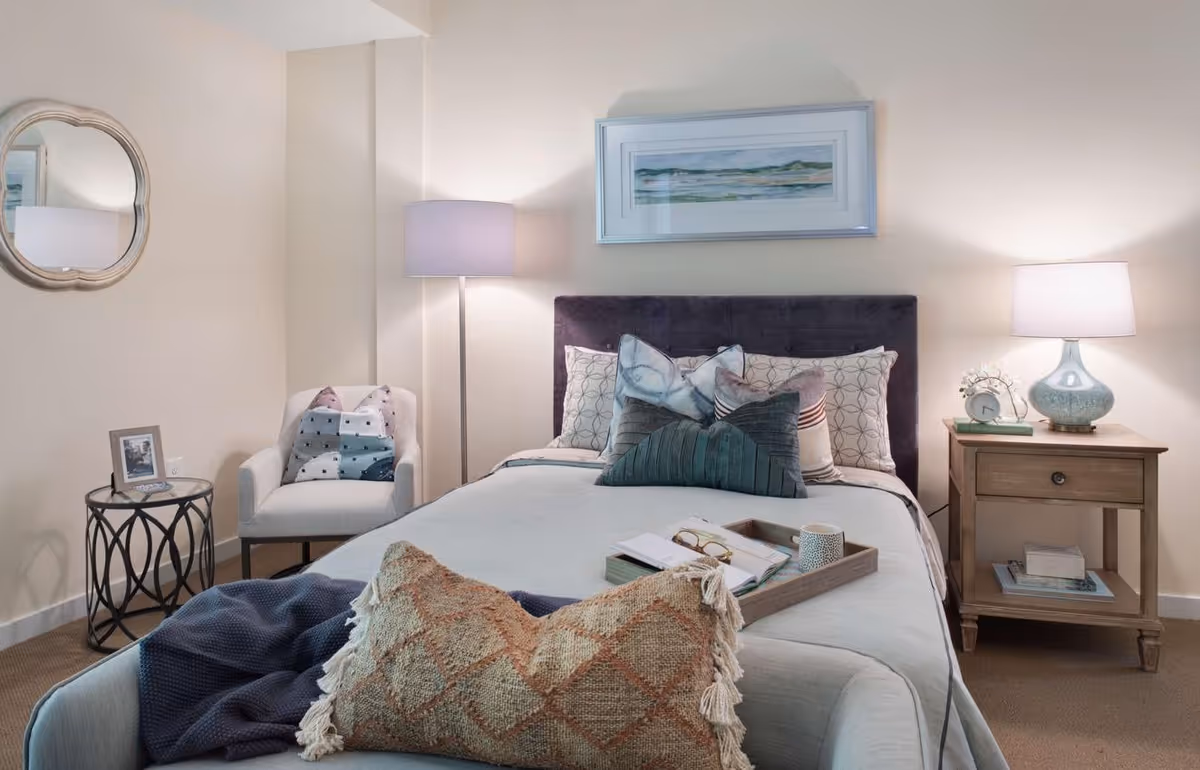 Cozy furnished bedroom with a made bed, decorative pillows, bedside tables with lamps, an armchair, and wall art.