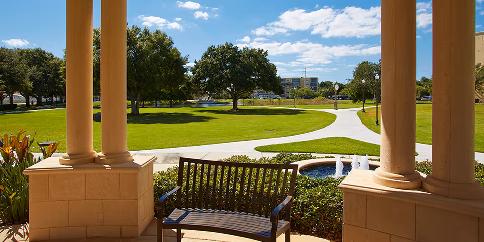 Stone-columned porch with a wooden bench overlooking a grassy courtyard, fountain, and walking paths.