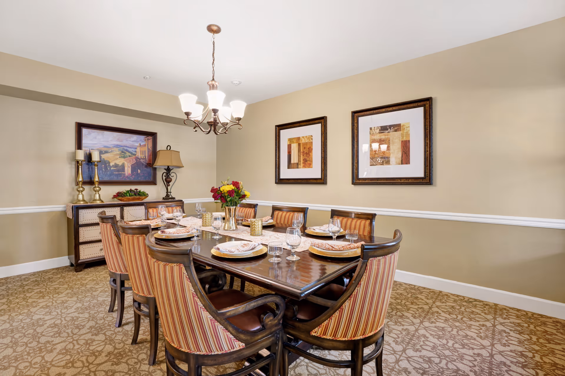 A dining room with a rectangular wooden table set for eight people. The chairs have striped upholstery. The table is set with plates, glasses, and napkins, and there is a vase with flowers in the center. The room has beige walls with white trim, two framed abstract paintings on one wall, and a sideboard with decorative items including candles and a lamp. A chandelier with five lights hangs above the table.