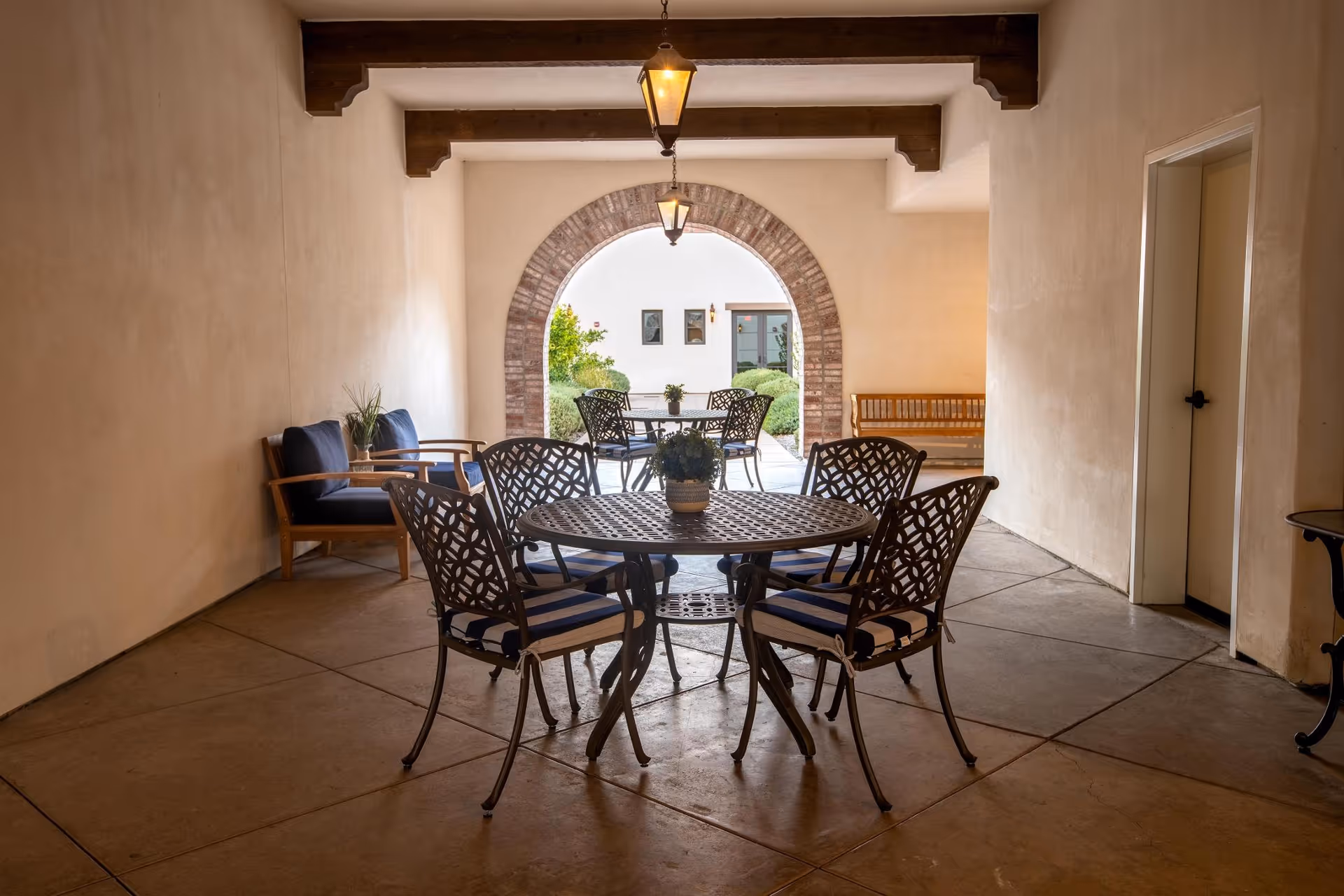Covered outdoor seating area with a round metal table and four chairs with blue and white striped cushions. There are two wooden armchairs with blue cushions against the left wall and a wooden bench on the right side near a doorway. The area opens to a courtyard through a brick archway, with additional tables and chairs visible outside. The space is lit by hanging lantern-style lights.