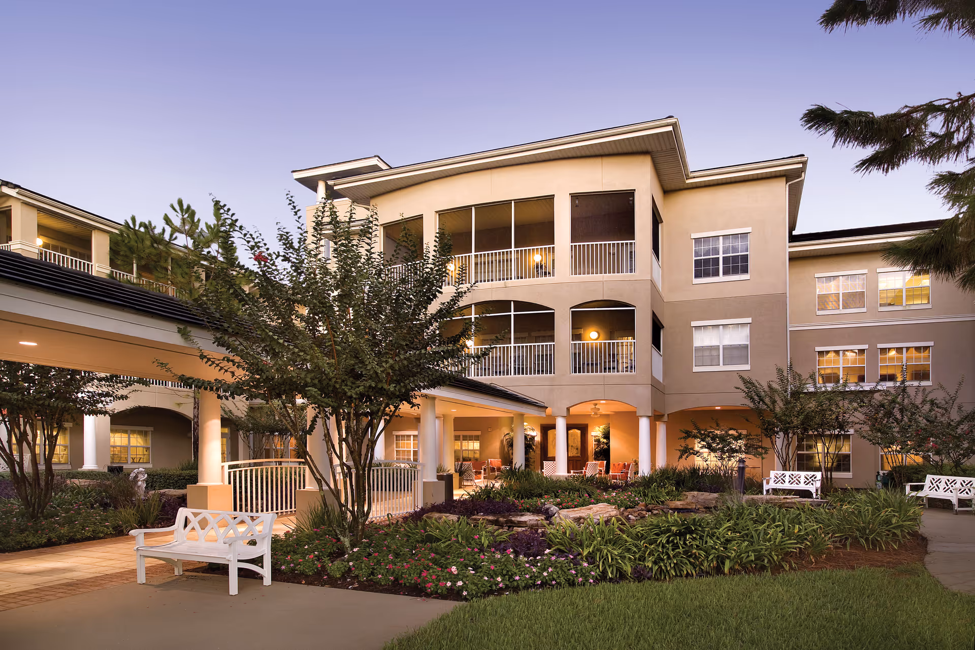 Exterior view of a senior living facility building with three stories, featuring balconies and large windows. The foreground shows a landscaped garden with green plants, flowers, trees, white benches, and a covered walkway leading to the entrance. The sky is clear with a soft evening light.
