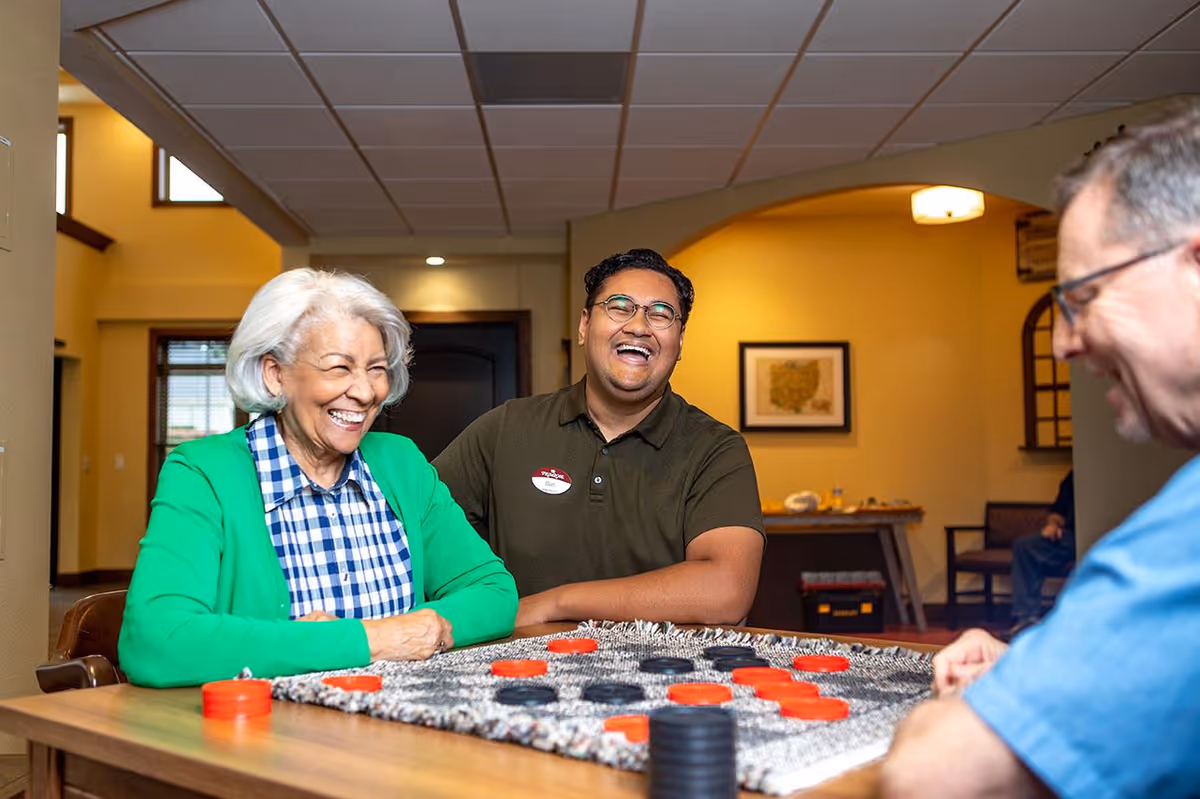 Three people sitting around a table playing a game of checkers in a warmly lit room. An elderly woman in a green cardigan and blue checkered shirt is smiling, a man in a dark green polo shirt with a name tag is laughing, and another man in a blue shirt is partially visible. The room has yellow walls, framed artwork, and wooden furniture.