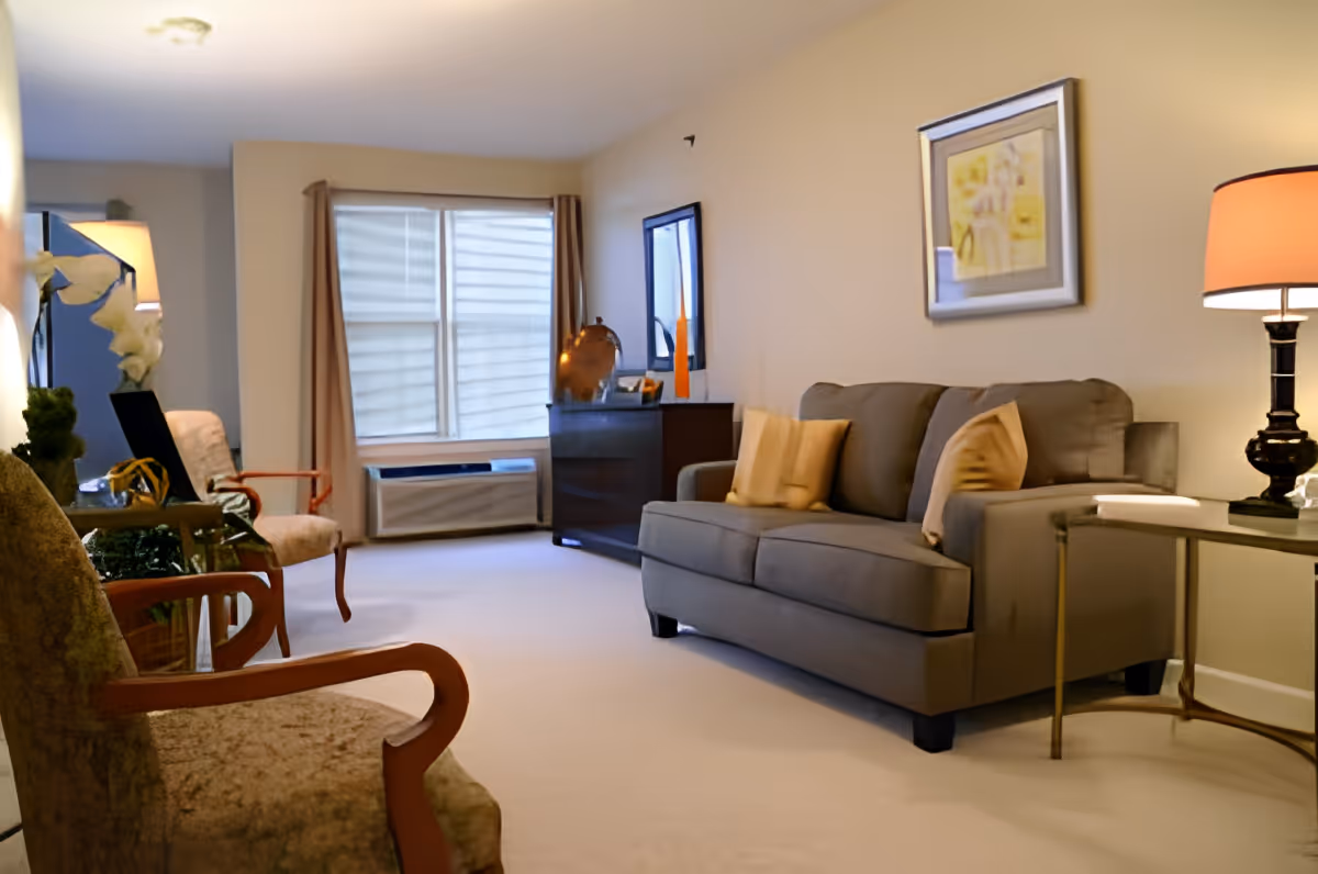 A cozy living room with a gray sofa adorned with beige pillows, two upholstered armchairs with wooden arms, a side table with a lamp, a window with beige curtains, and a dark wooden cabinet with decorative items.