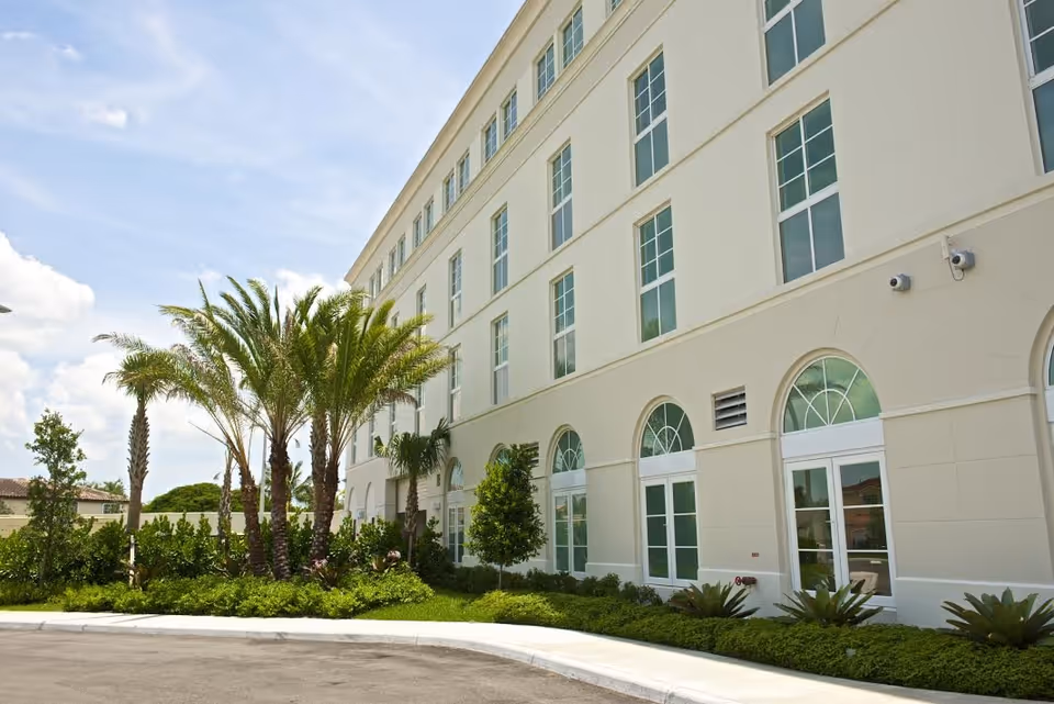 Three-story beige senior living facility with arched ground-floor windows and palm-lined entrance.