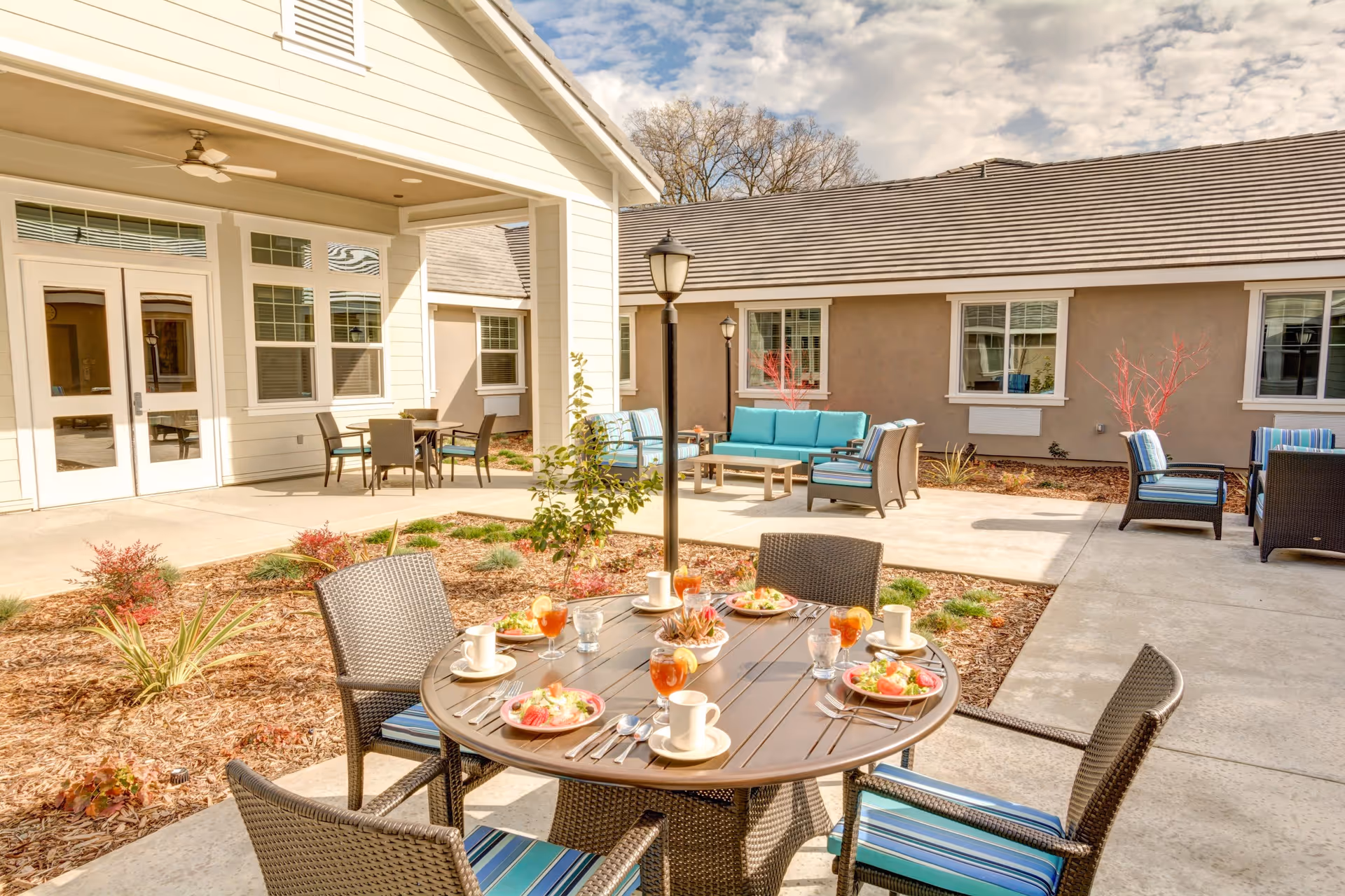 Outdoor patio area at The Courte At Citrus Heights with a round table set for four with plates of food, glasses of iced tea, and coffee cups. Surrounding the table are wicker chairs with blue striped cushions. In the background, there are additional seating areas with blue cushioned sofas and chairs, beige building walls with windows, and a clear sky with some clouds.