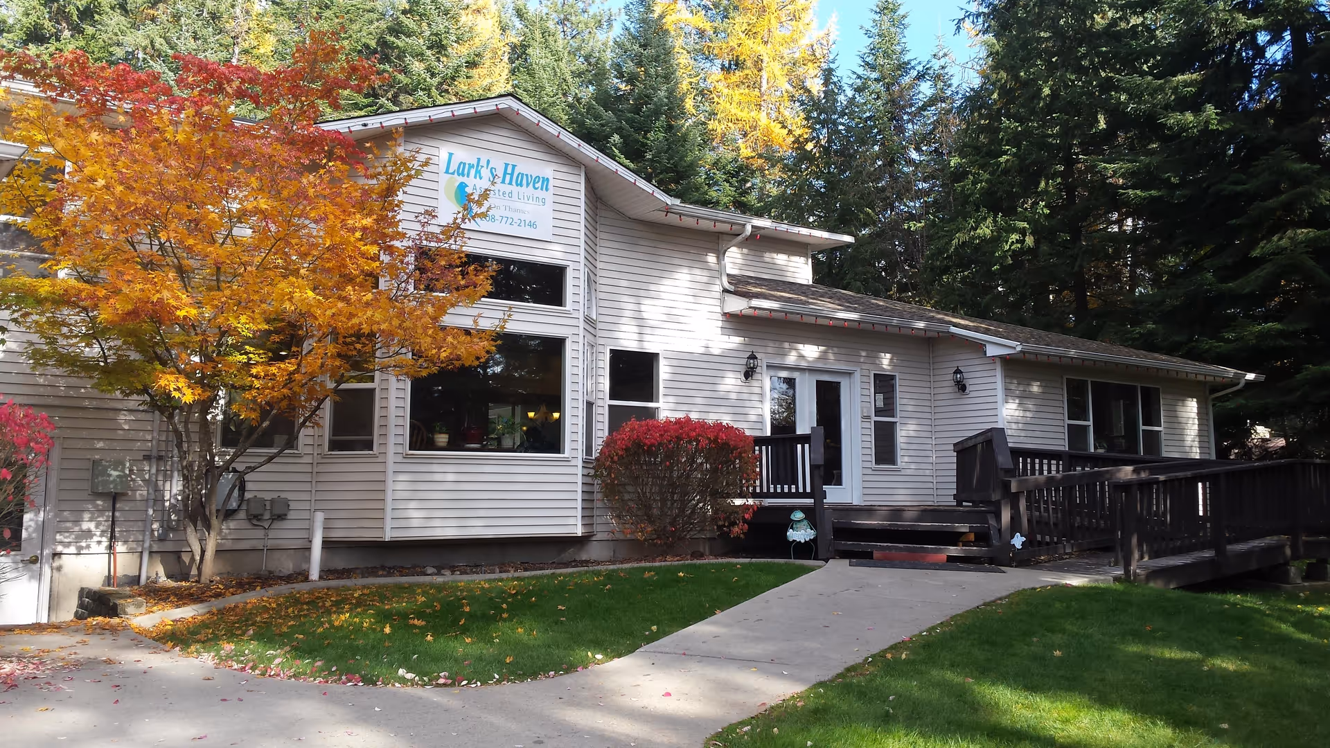 Exterior view of Larkshaven Assisted Living facility showing a single-story building with beige siding, large windows, a wheelchair-accessible ramp, and a concrete walkway. There are colorful autumn trees and green grass surrounding the building, with tall evergreen trees in the background.