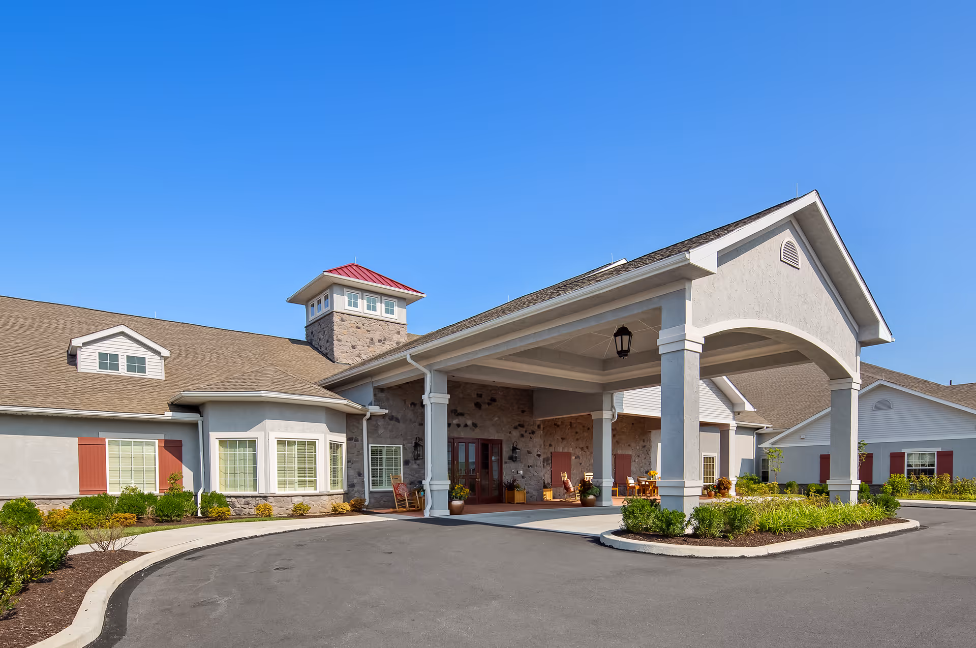 Exterior view of the Legend of Allentown senior living facility showing the main entrance with a covered drop-off area, stone and siding facade, red shutters, and a clear blue sky.