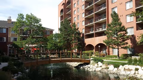 Outdoor courtyard featuring a wooden footbridge over a pond, trees, and a brick residential building.
