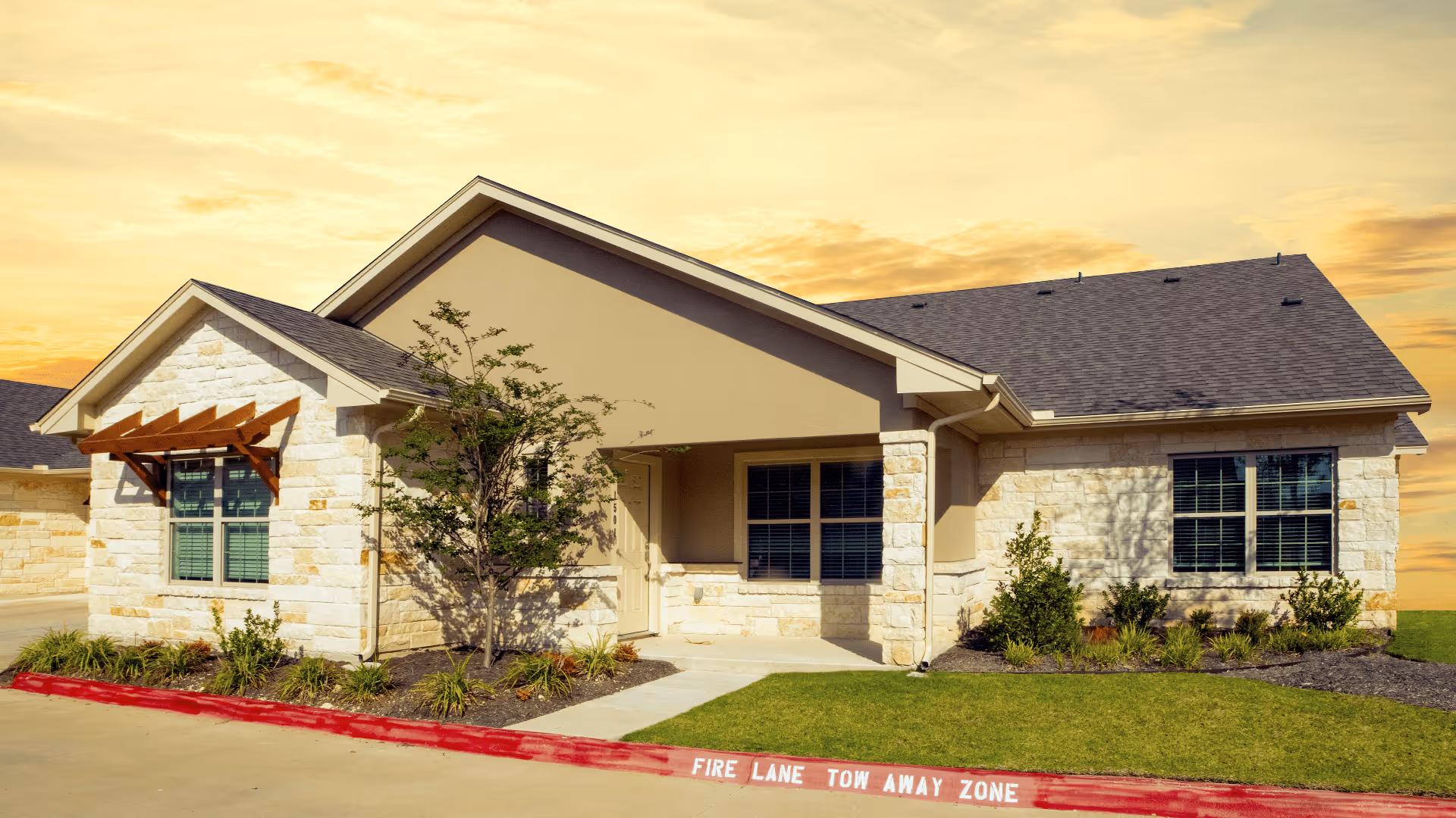 Exterior view of a single-story residential building with beige stone walls and a dark shingled roof under a yellow-orange sky at sunset. The building has multiple windows with blinds, a small covered entrance, and landscaped greenery including bushes and a tree. A red curb with white text reading 'FIRE LANE TOW AWAY ZONE' is visible in front of the building.