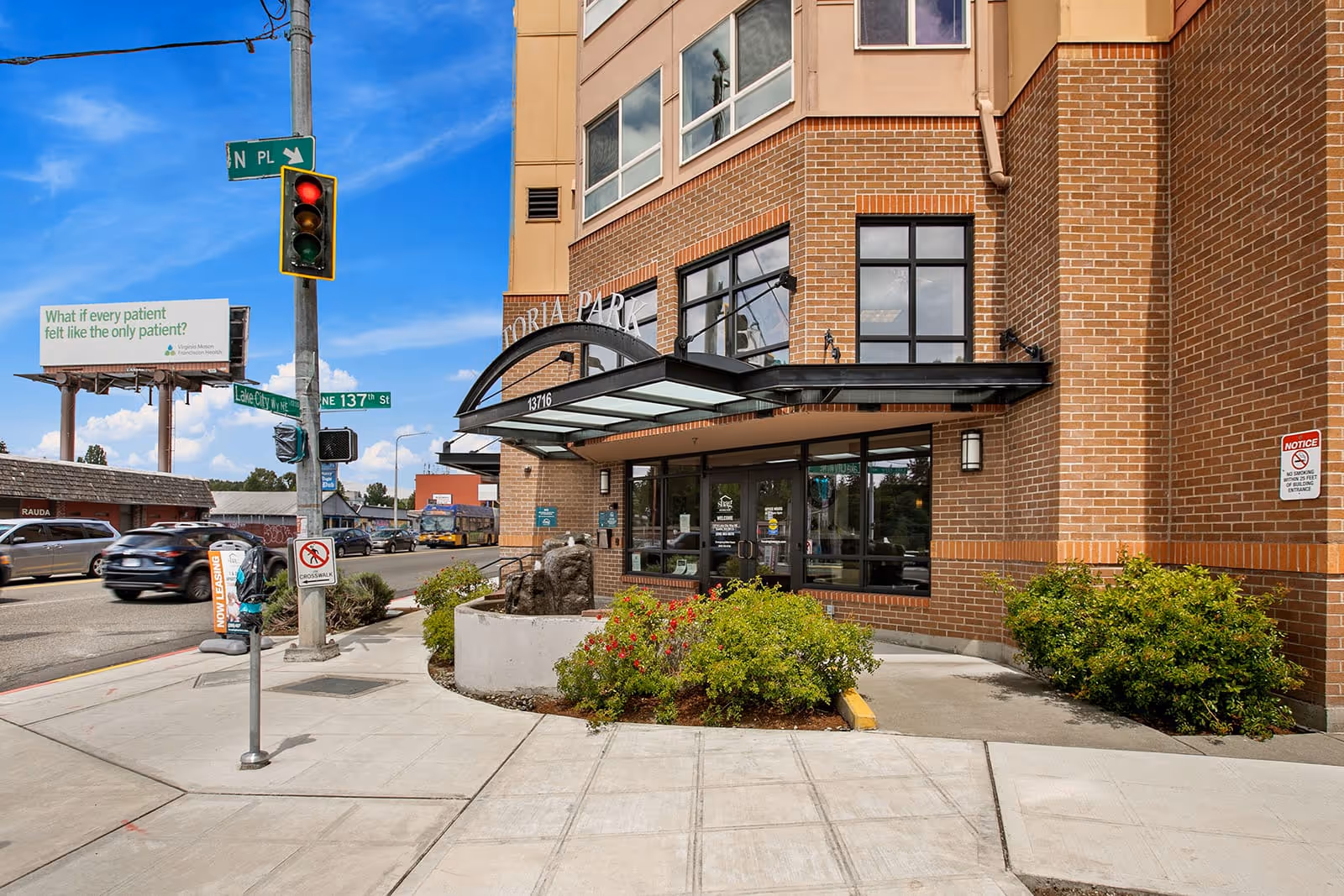 Entrance of Victoria Park Senior Living showing a brick facade and glass awning at a street corner with sidewalk and traffic light.