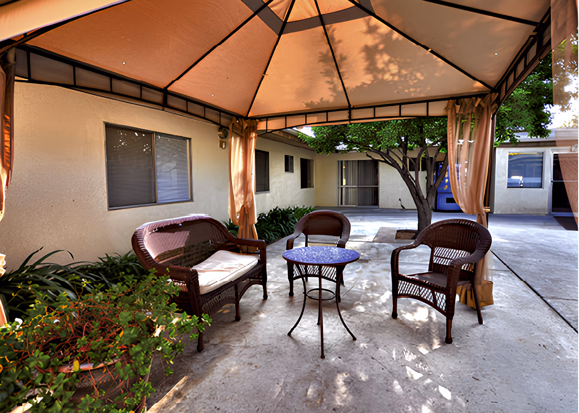 Outdoor seating area under a canopy with a small round table, two chairs, and a loveseat with cushions, surrounded by plants and trees near a building with windows and doors.