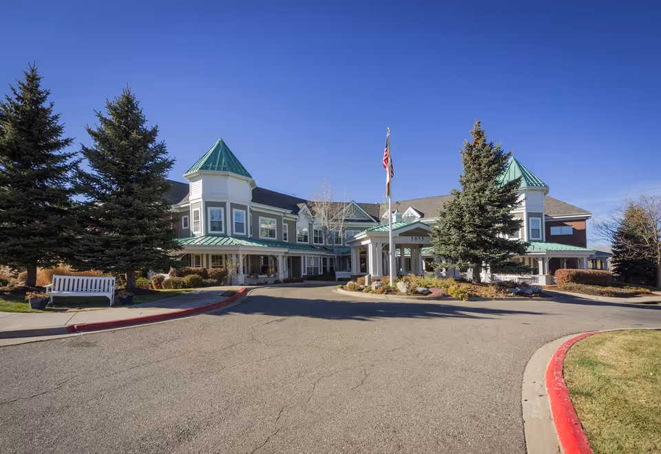Exterior view of a senior living facility building with green roofs and white trim, surrounded by evergreen trees and landscaping under a clear blue sky. There is a circular driveway with a flagpole in front of the entrance.