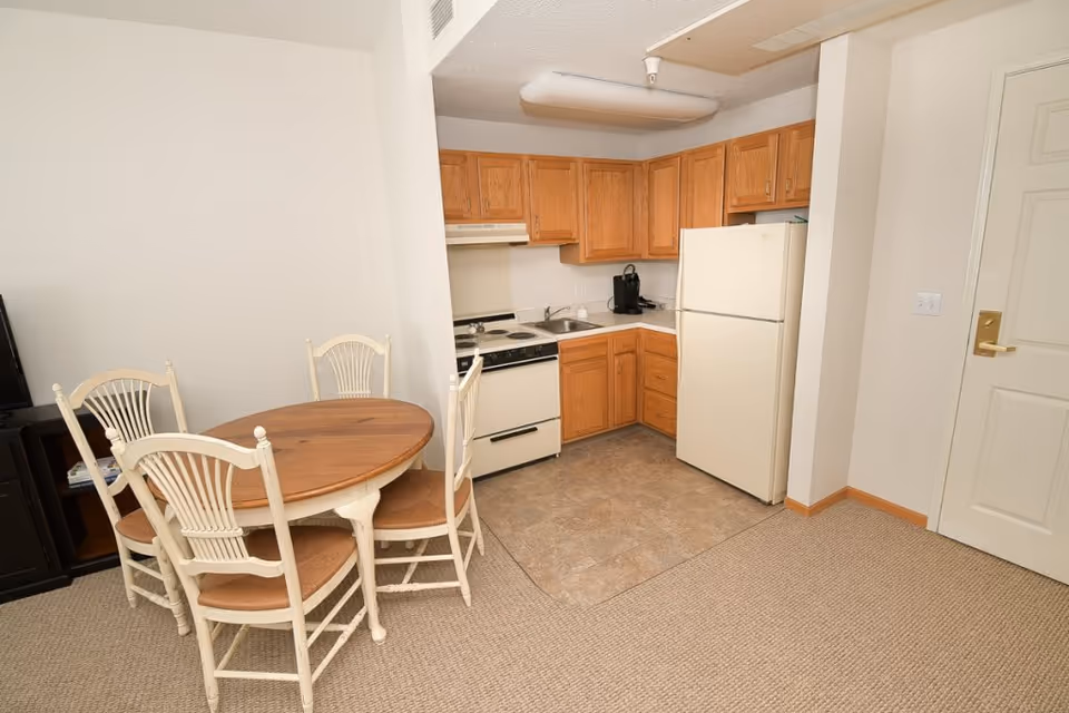 Small kitchen area with wooden cabinets, a white refrigerator, stove, and sink. Adjacent to the kitchen is a round wooden dining table with four white chairs. The floor transitions from tile in the kitchen area to carpet in the dining area. A white door with a gold handle is visible on the right side.