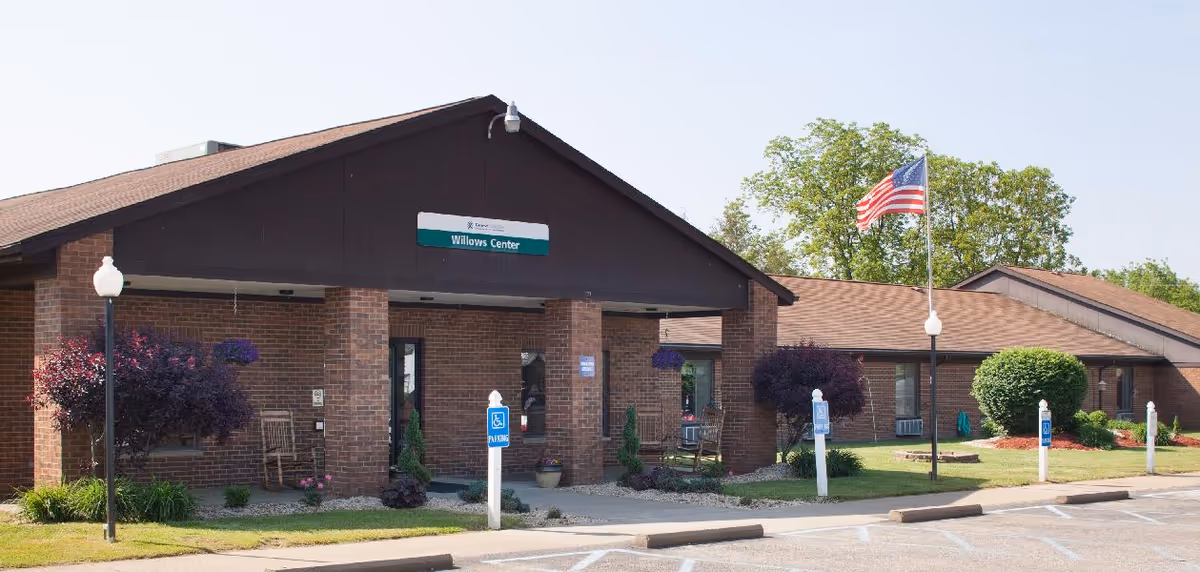 Exterior view of the Willows Center building, a single-story brick structure with a brown roof. The entrance has a covered porch supported by brick columns, with rocking chairs on the porch. There are several handicap parking signs and spaces in front of the building. An American flag is flying on a flagpole near the entrance, and there are well-maintained bushes and trees around the building.