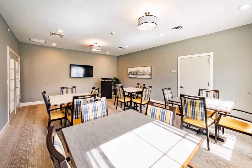 Bright communal dining room with several square tables and plaid-backed chairs, a wall-mounted TV and artwork on muted green walls.