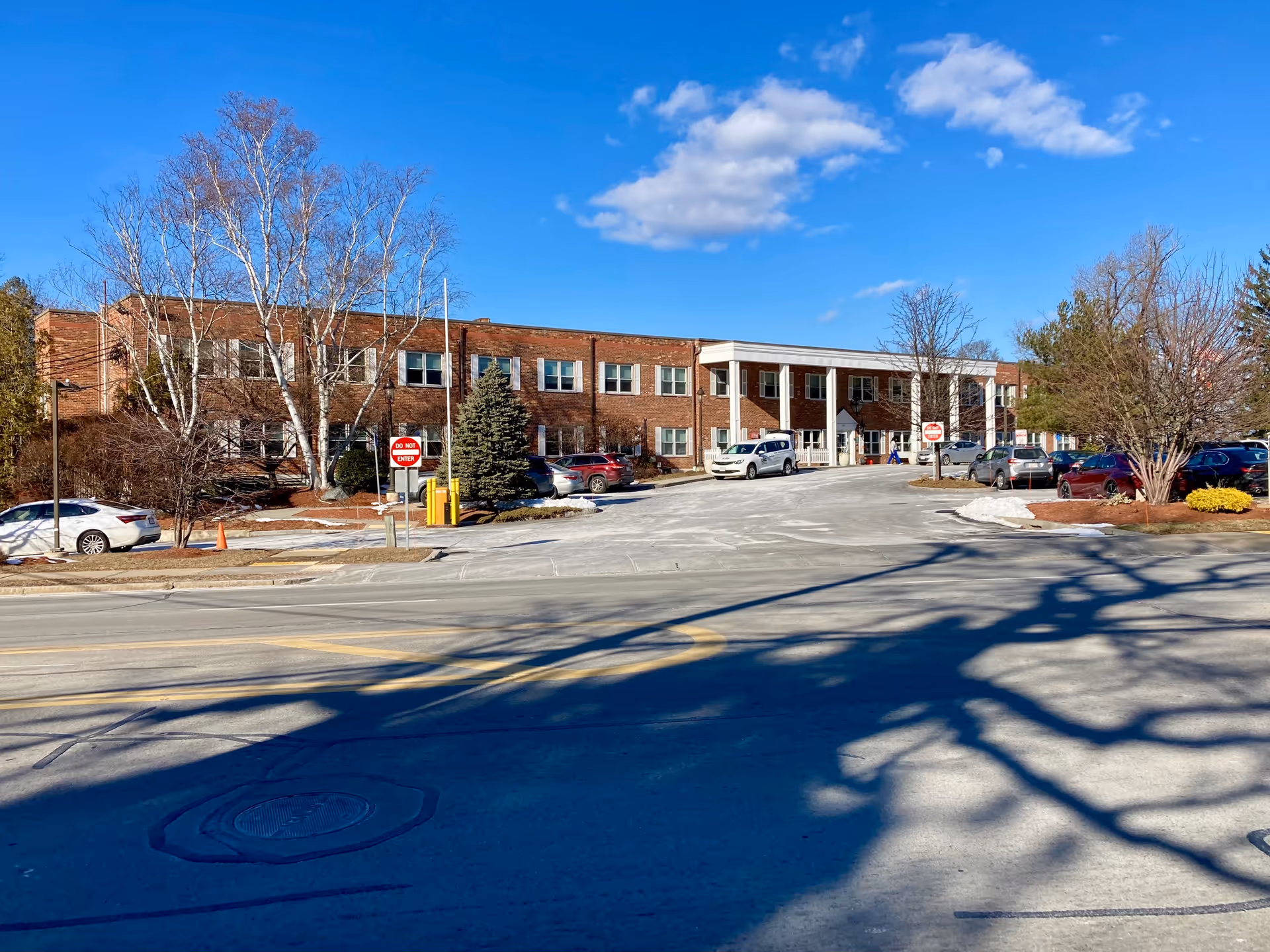 Exterior view of a two-story brick building with multiple windows, a driveway with parked cars, leafless trees, and a clear blue sky with some clouds.